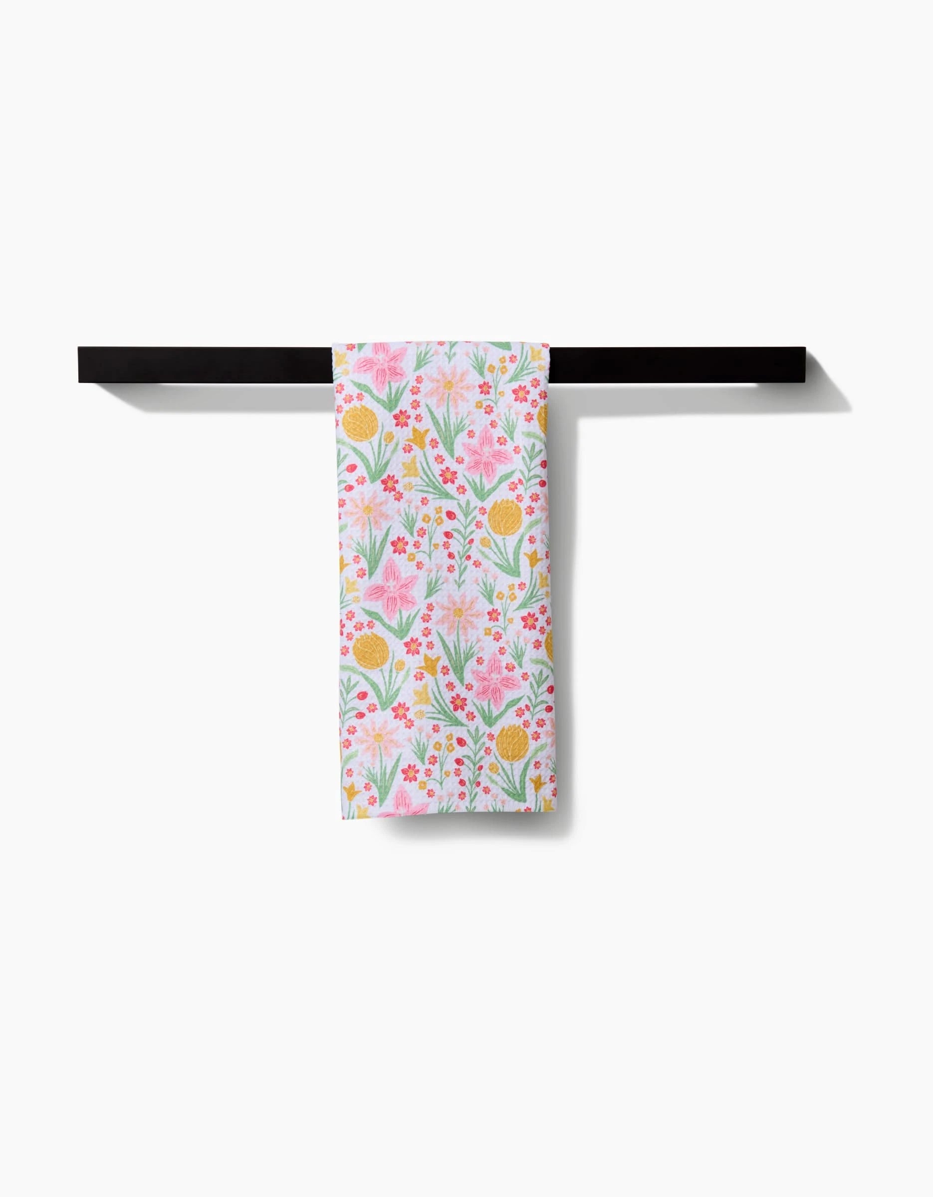 The Garden of Light tea towel by Danelys Sidron, featuring a vibrant pink, yellow, and green floral pattern, is neatly draped over a black horizontal bar against a white background.