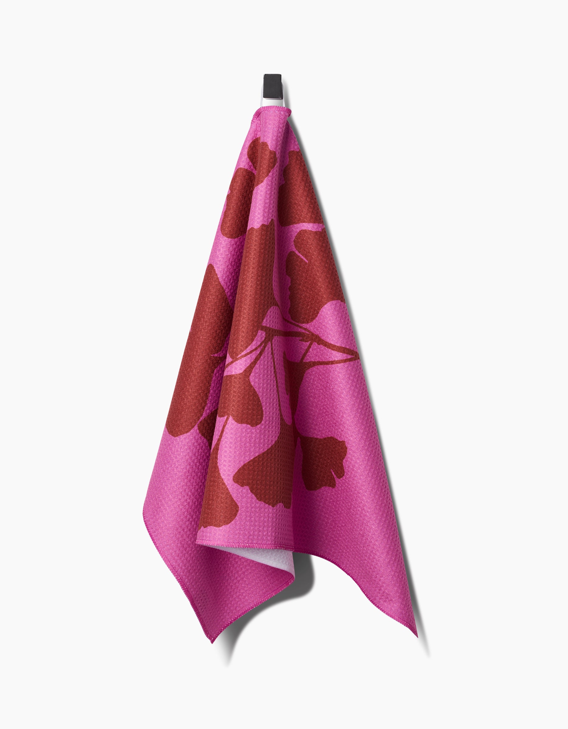 PrintedPlant’s Ginkgo dish towel, in pink with a bold red abstract floral pattern and made from ultra-absorbent Geoweave fabric, hangs on a black hook against a plain white background.