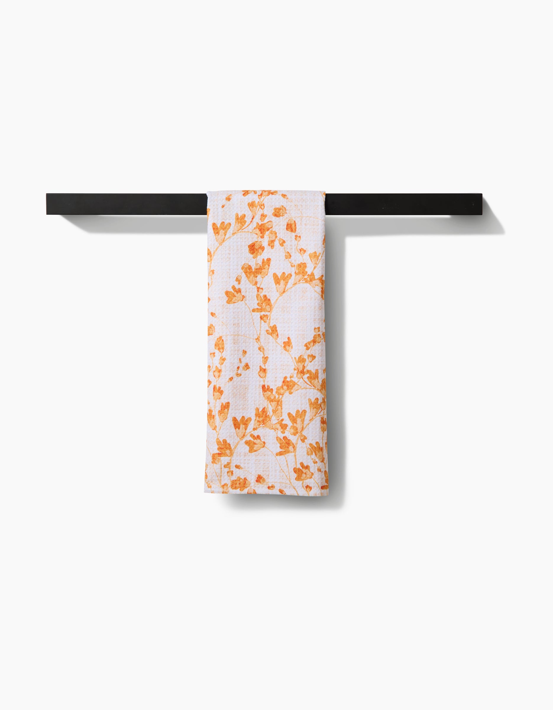 The Pascale Dilger Golden Grassland Tea Towel, featuring an orange floral pattern, hangs on a black bar against a white background. Made from Geoweave™ fabric, it offers superior absorbency and performance.
