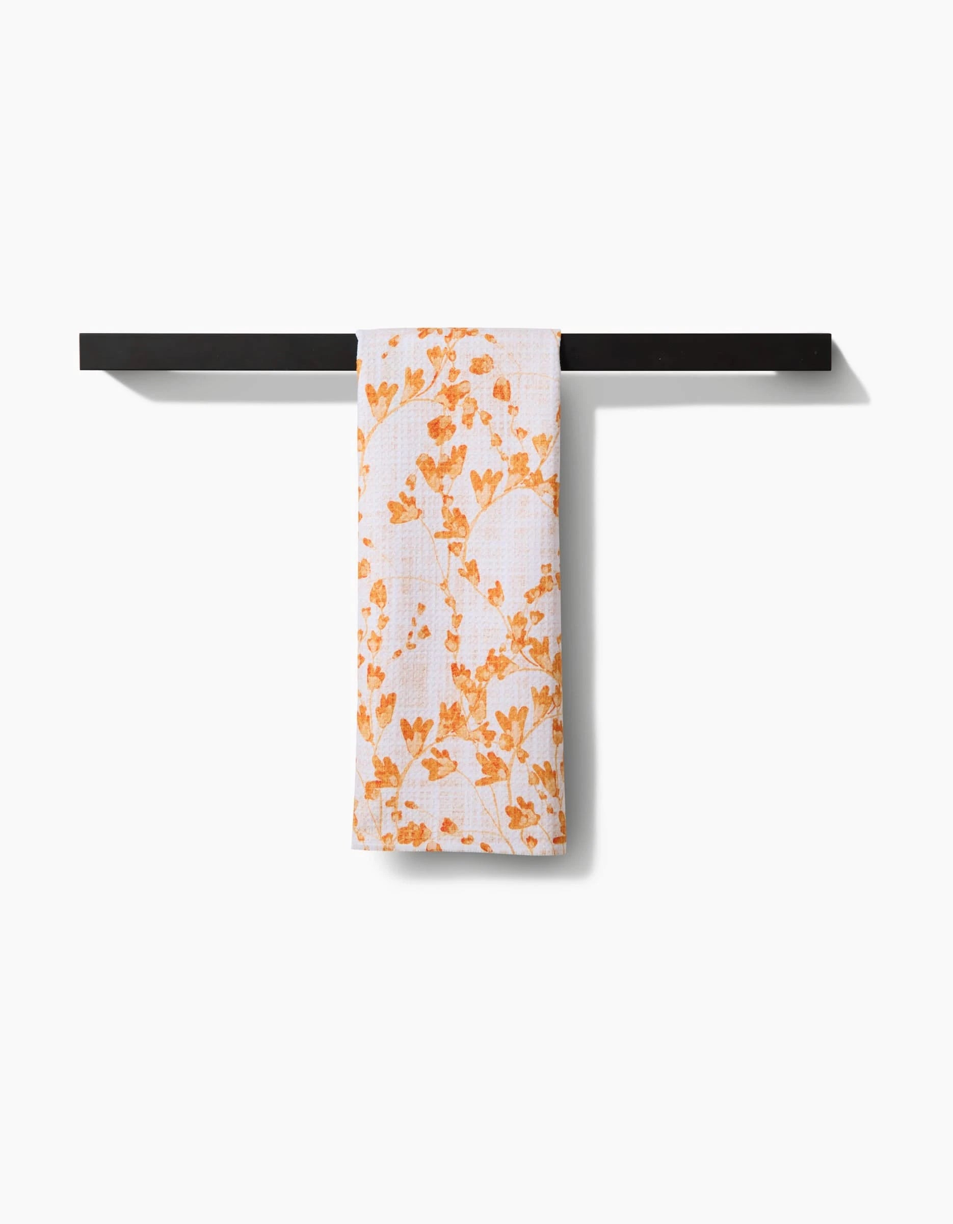 The Pascale Dilger Golden Grassland Tea Towel, featuring an orange floral pattern, hangs on a black bar against a white background. Made from Geoweave™ fabric, it offers superior absorbency and performance.