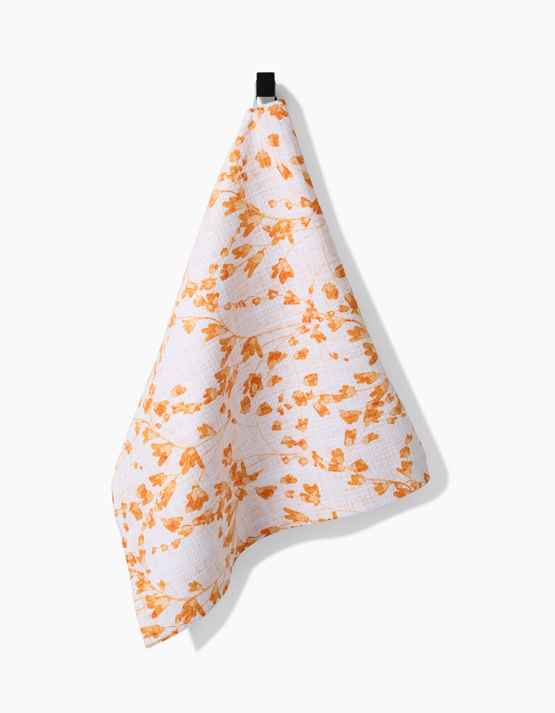 The Pascale Dilger Golden Grassland tea towel, made from white Geoweave™ fabric with an orange floral pattern, hangs from a black loop against a plain white background.