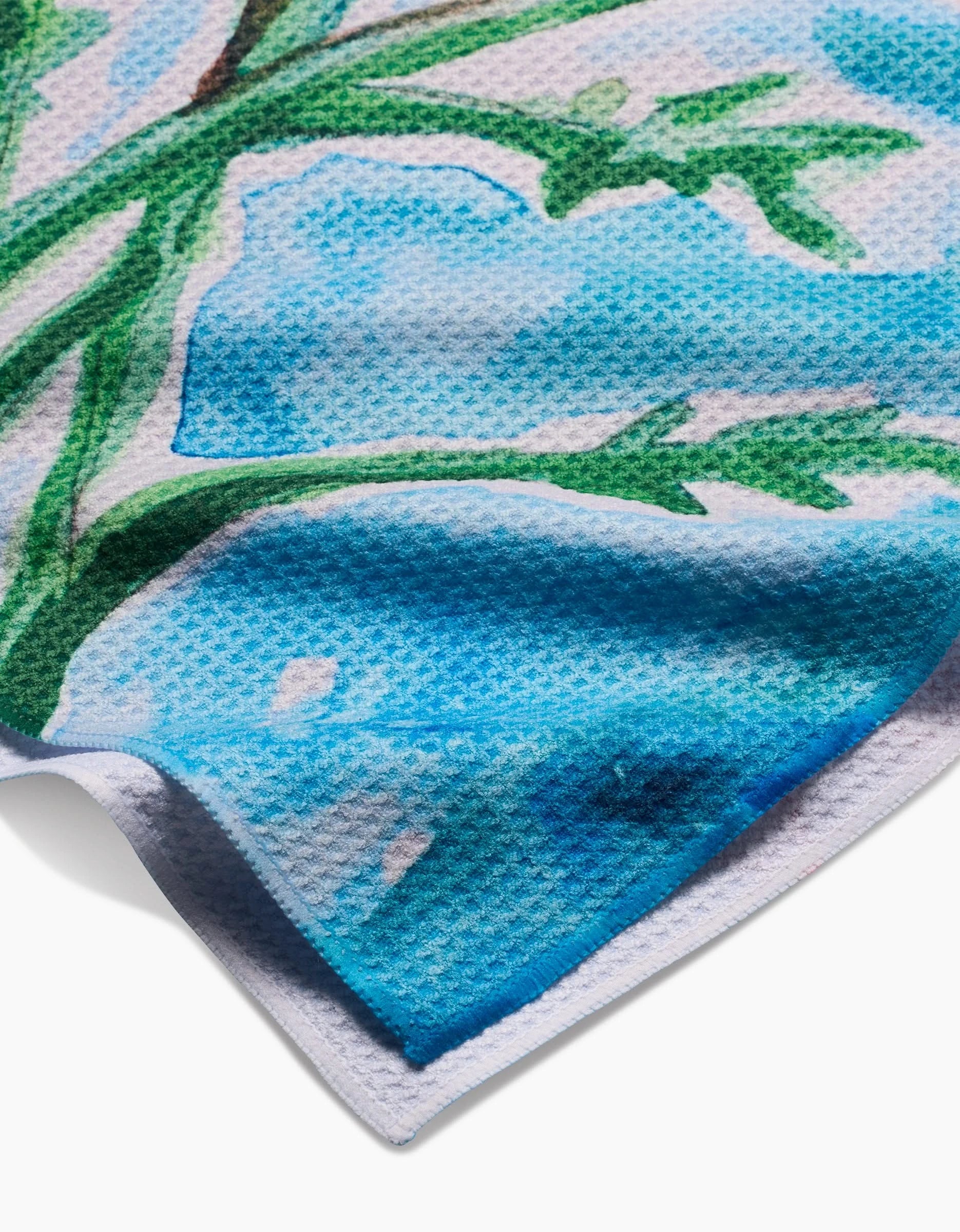 A close-up of Skye Walker’s Golden Poppy Flowers Geoweave™ tea towel, featuring a blue, green, and white watercolor floral pattern with the corner slightly folded.