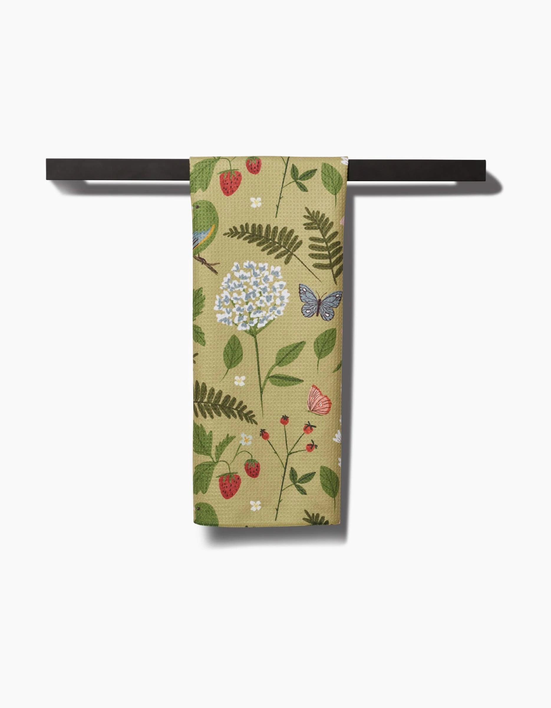 The Good Green Earth tea towel by Janna Sue Design features a beige background with white flowers, green leaves, strawberries, and butterflies. Made from recycled materials, it hangs on a black towel bar against a white backdrop.