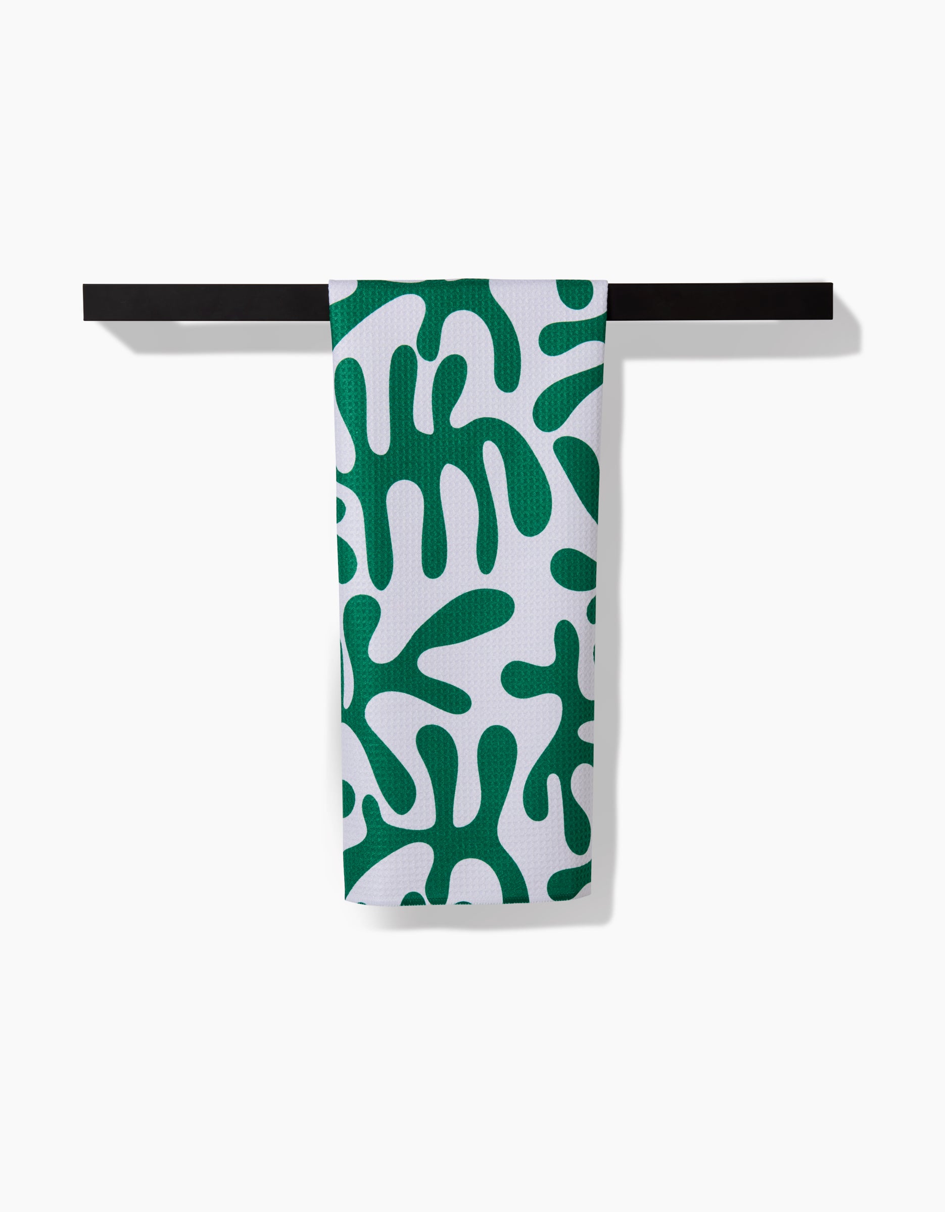 A Green Afternoon towel by Marina Solodka, featuring bold green abstract leaf patterns on white ultra-absorbent Geoweave fabric, is draped over a simple black rod against a plain white background.