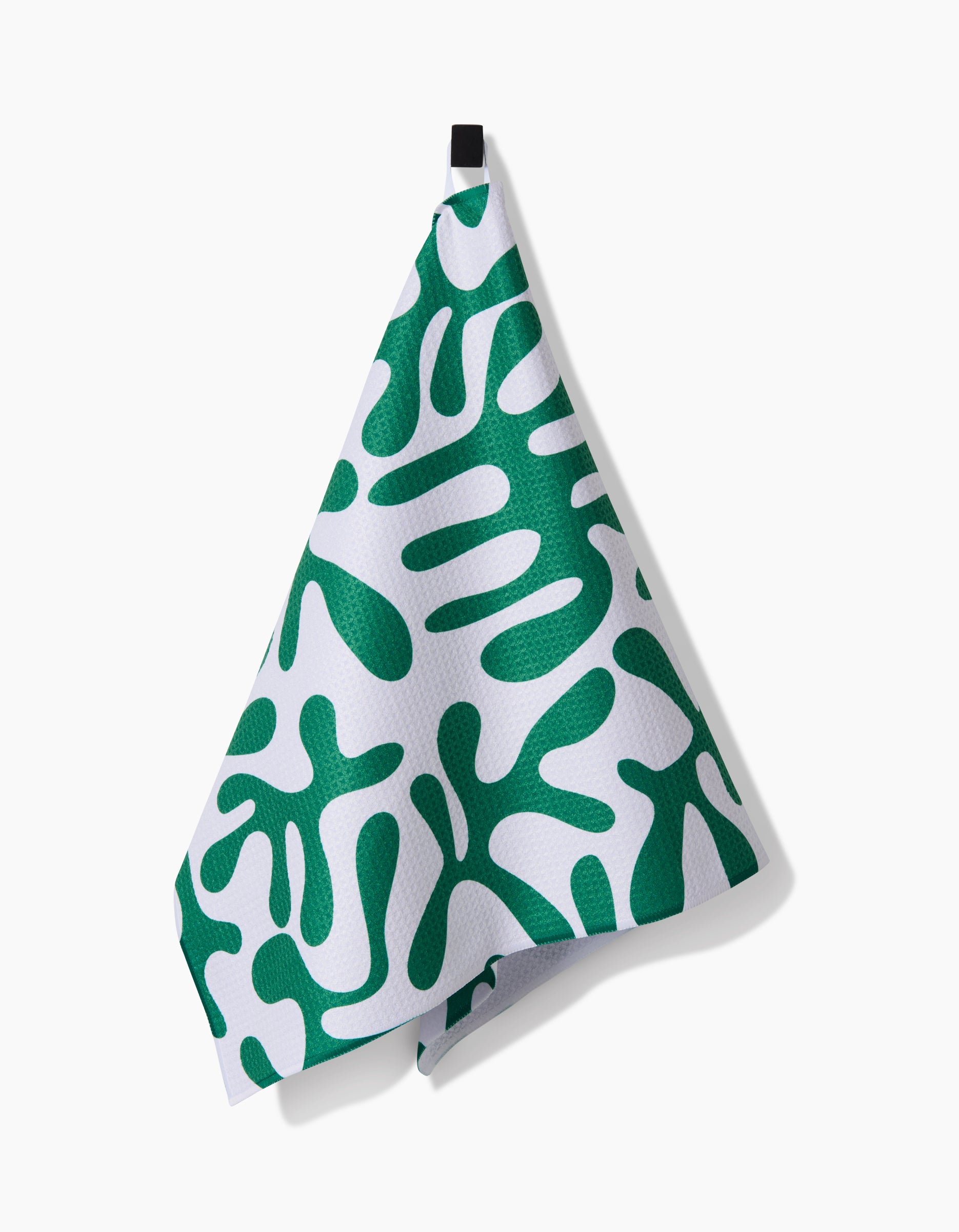 The Green Afternoon towel by Marina Solodka, made from ultra-absorbent Geoweave fabric, features bold green abstract organic shapes on white and hangs from a black loop against a white background.