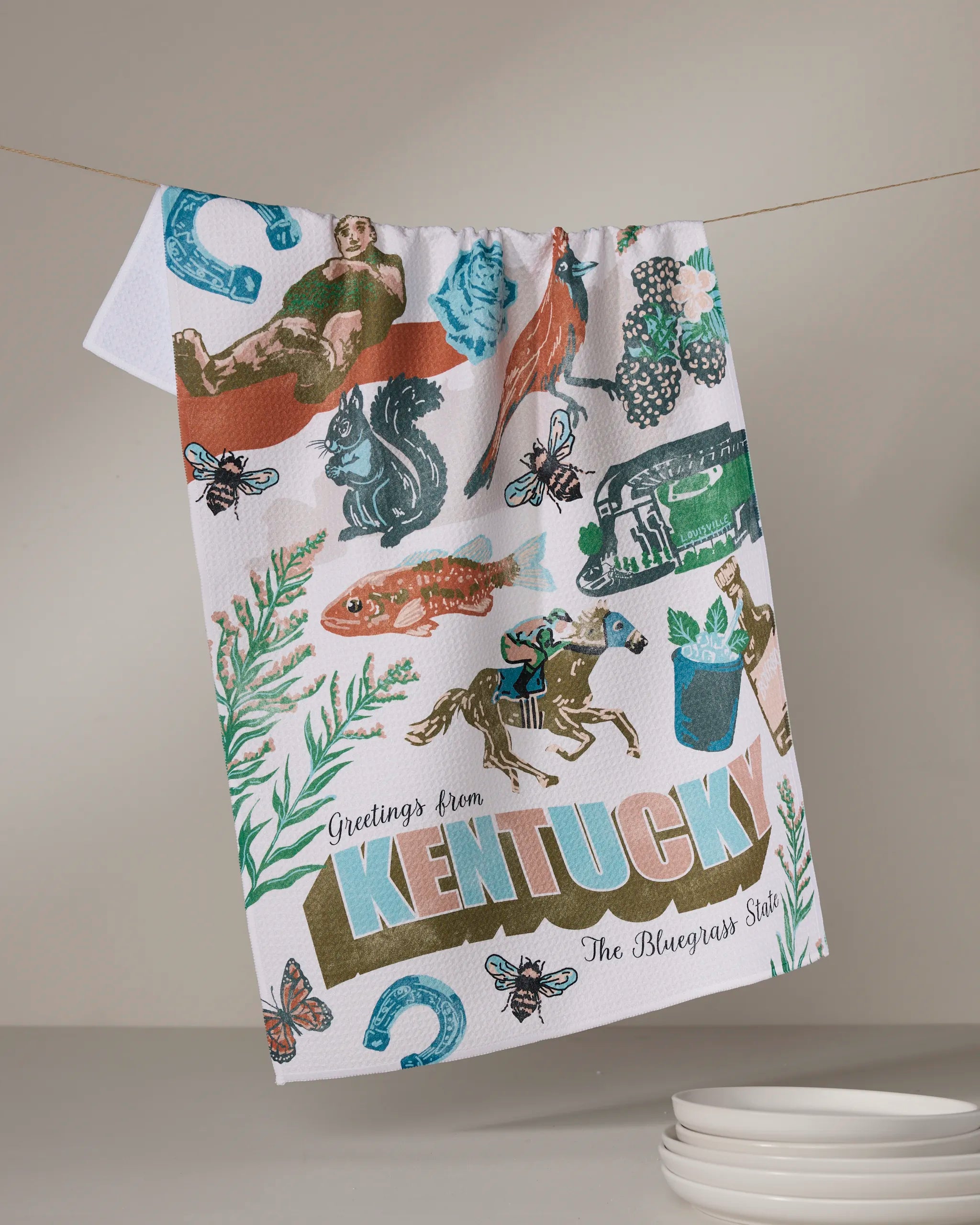 A recycled Geometry kitchen towel, “Greetings From Kentucky,” hangs on a line, featuring colorful prints of a horse, jockey, fish, flowers, bees, horseshoes, and “Greetings from KENTUCKY The Bluegrass State.”.