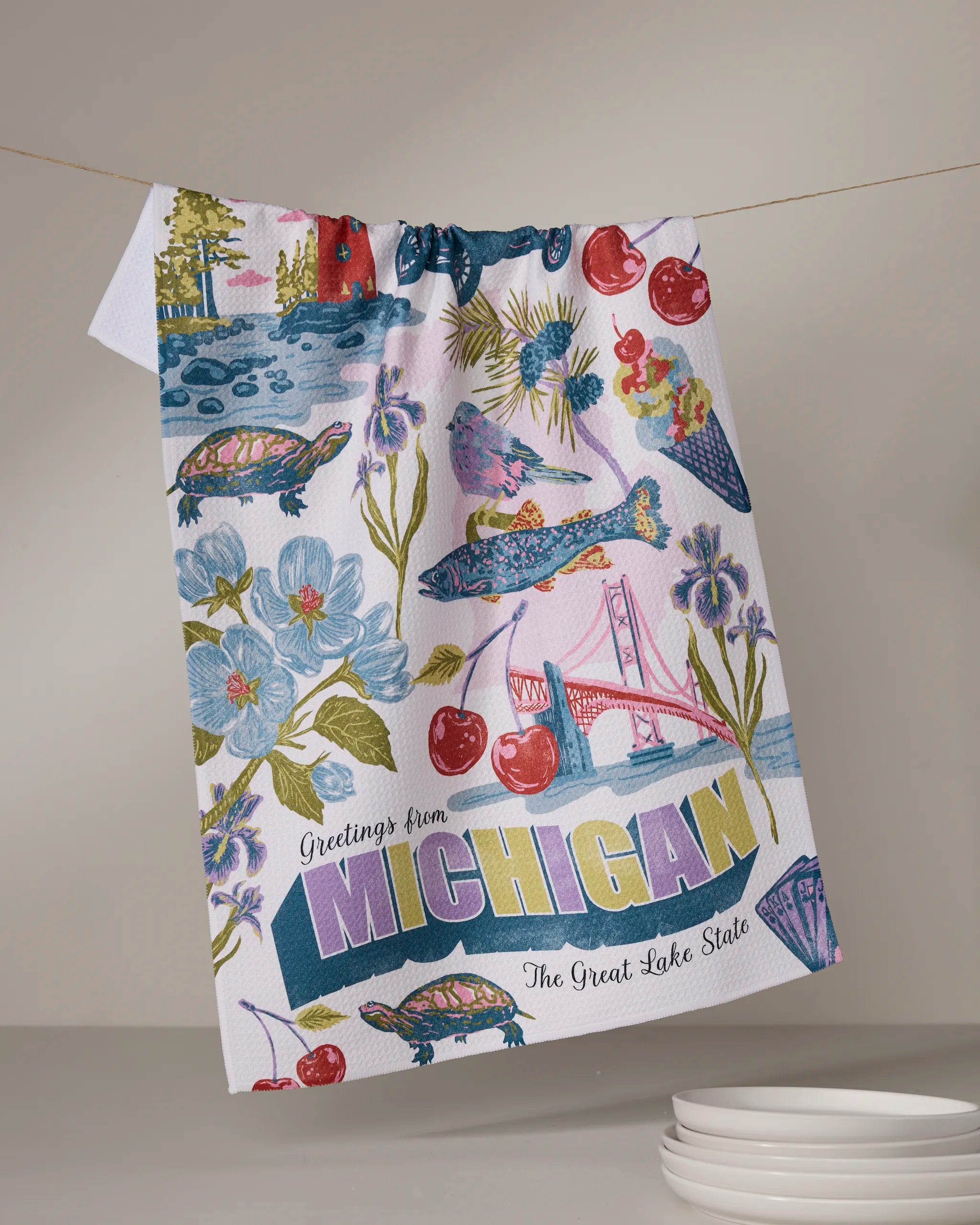 The Geometry "Greetings From Michigan" tea towel hangs on a line, ultra-absorbent and colorful, featuring Michigan landmarks, wildlife, cherries, ice cream, and the text "Greetings from Michigan, The Great Lake State.