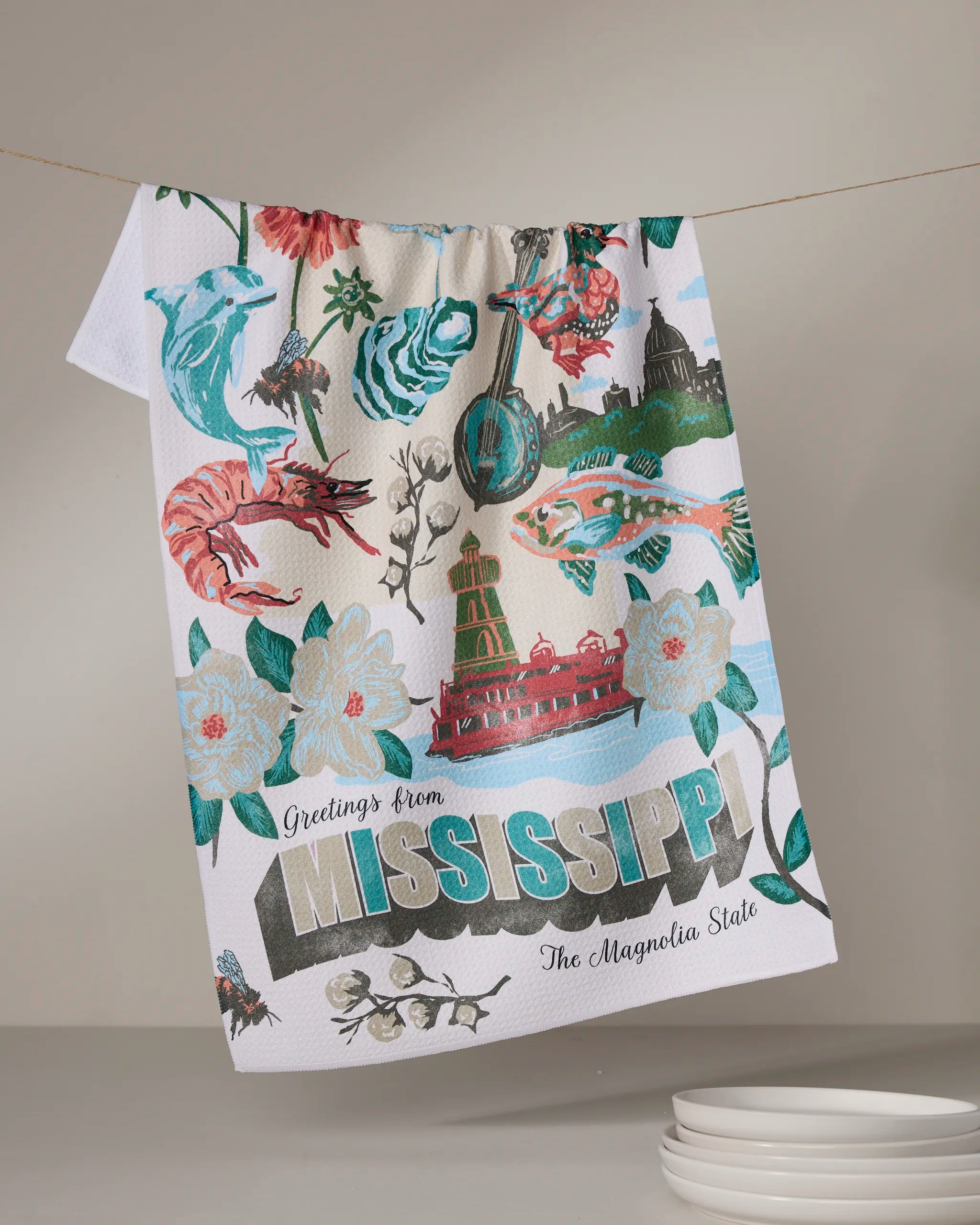 The Geometry "Greetings From Mississippi" tea towel features vibrant Magnolia State symbols—magnolias, shrimp, and a riverboat. Ultra-absorbent, eco-friendly, and made from recycled materials.