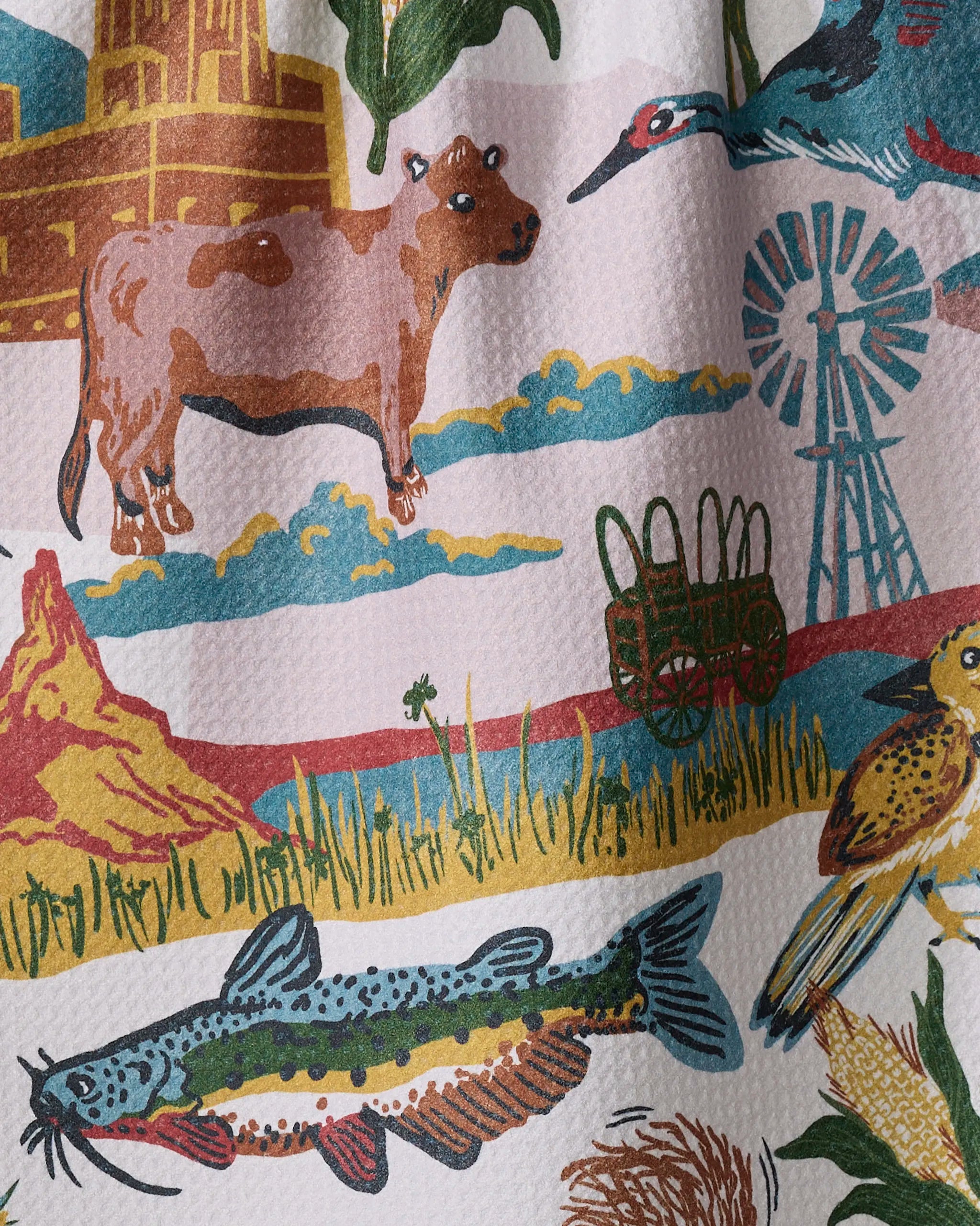 Brighten your kitchen with Geometry's "Greetings From Nebraska" tea towel—an eco-friendly design featuring a cow, windmill, covered wagon, catfish, bird, and yellow building among hills and grass under a blue sky.