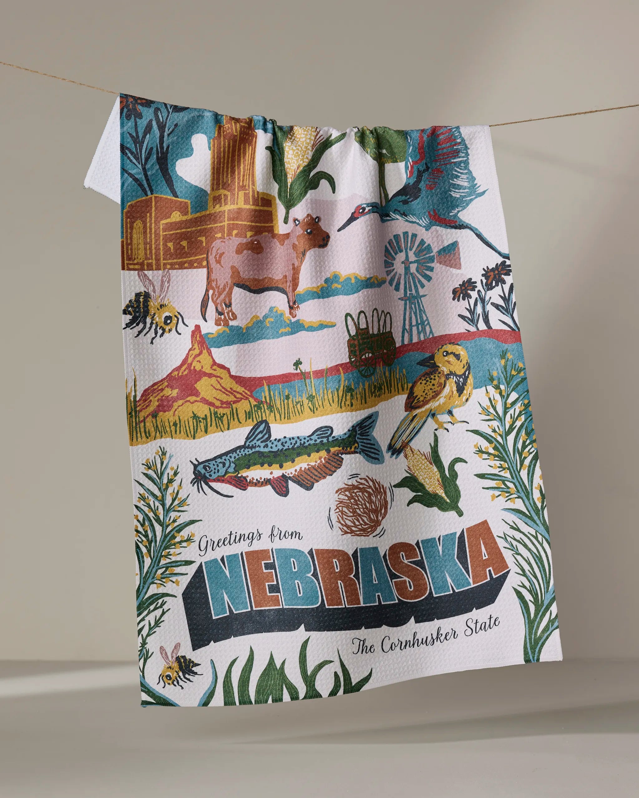 The Geometry "Greetings From Nebraska" kitchen towel showcases Nebraska’s wildlife, landmarks, and farmland with the text "Greetings from Nebraska, The Cornhusker State." Made from recycled materials for a sustainable touch.