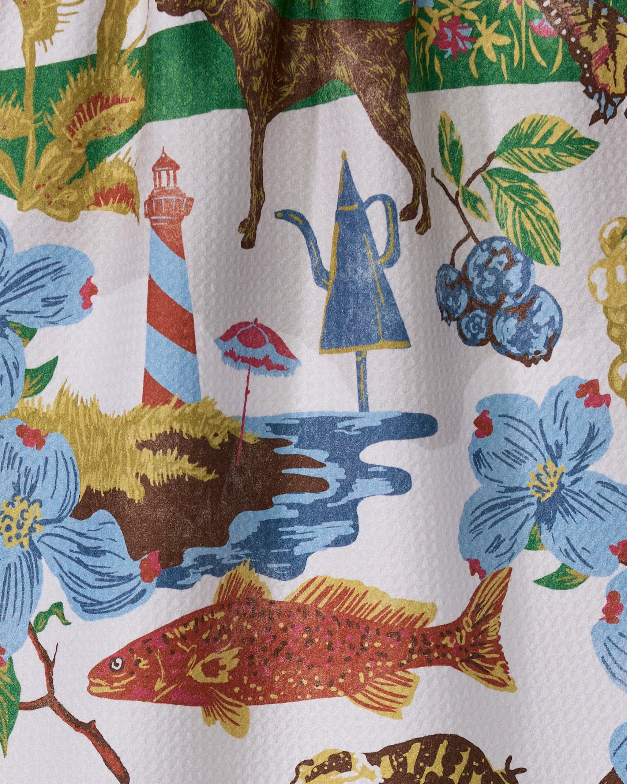 The "Greetings From North Carolina" tea towel by Geometry features a red fish, blue flowers, blueberries, a striped lighthouse, brown dog, shore umbrella, and teapot-shaped structure on recycled material.