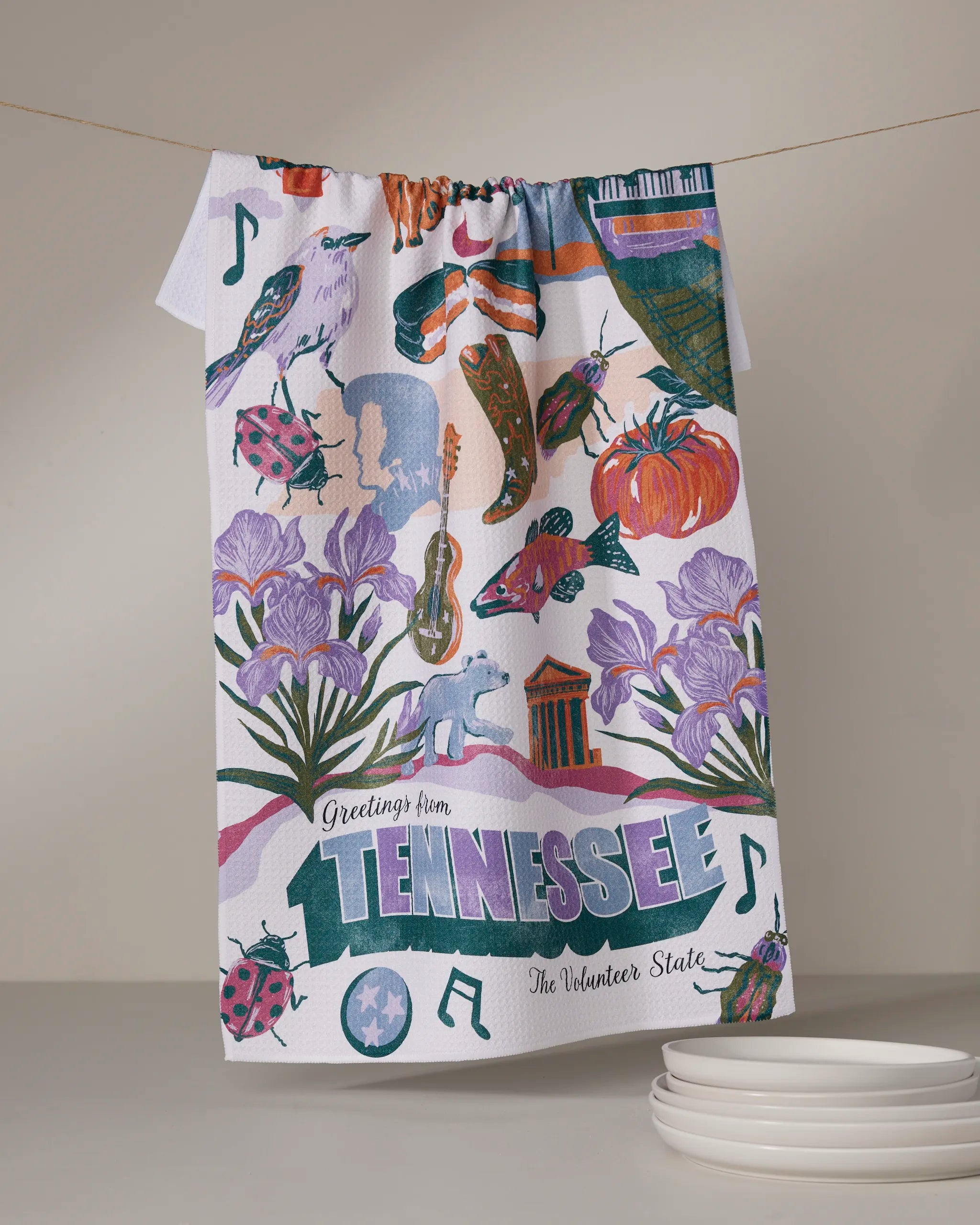 The Geometry "Greetings From Tennessee" tea towel, featuring vibrant state icons and phrases, hangs on a line above plates resting on the table.