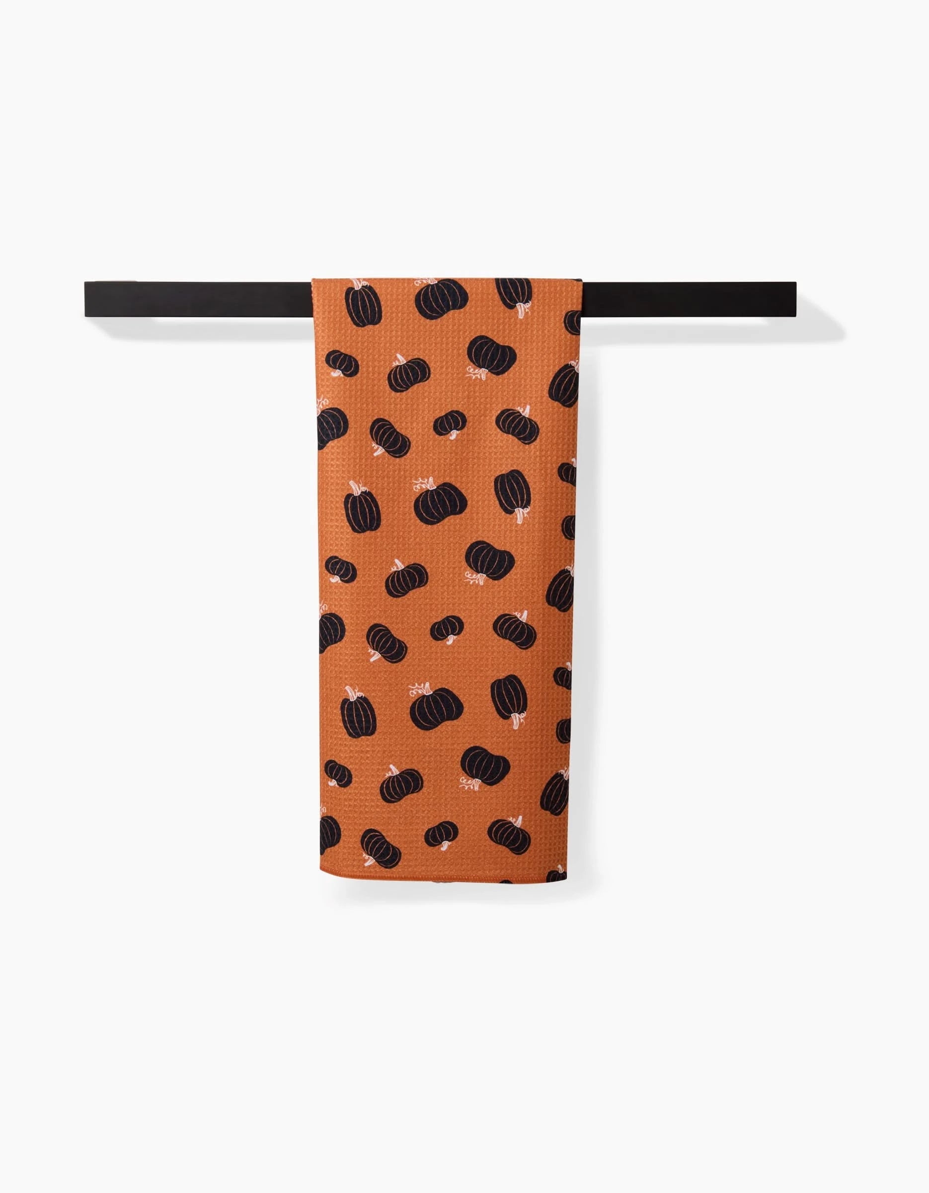 The Halloween Black Pumpkins tea towel by Hufton Studio, featuring a rust color and black pumpkin pattern, is neatly folded over a black rod against a white background.