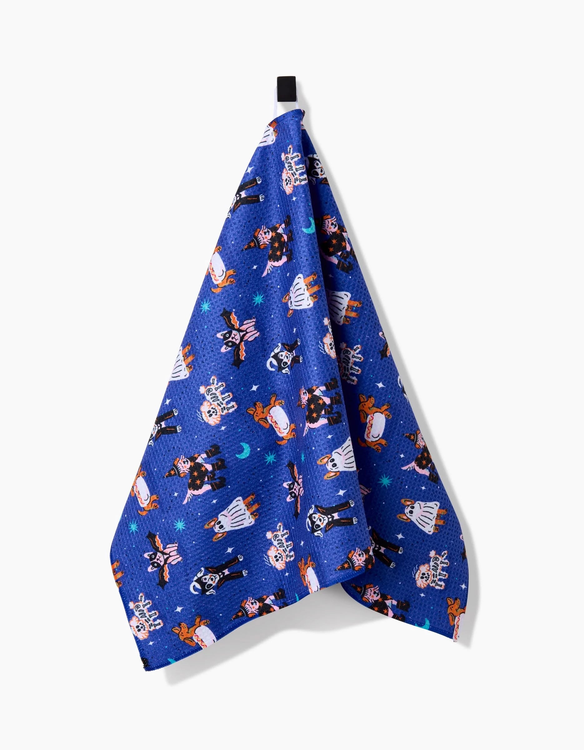 The Halloween Hounds tea towel by Camila Leão features playful dogs in costumes, crafted from recycled materials, and hangs on a hook against a white background.