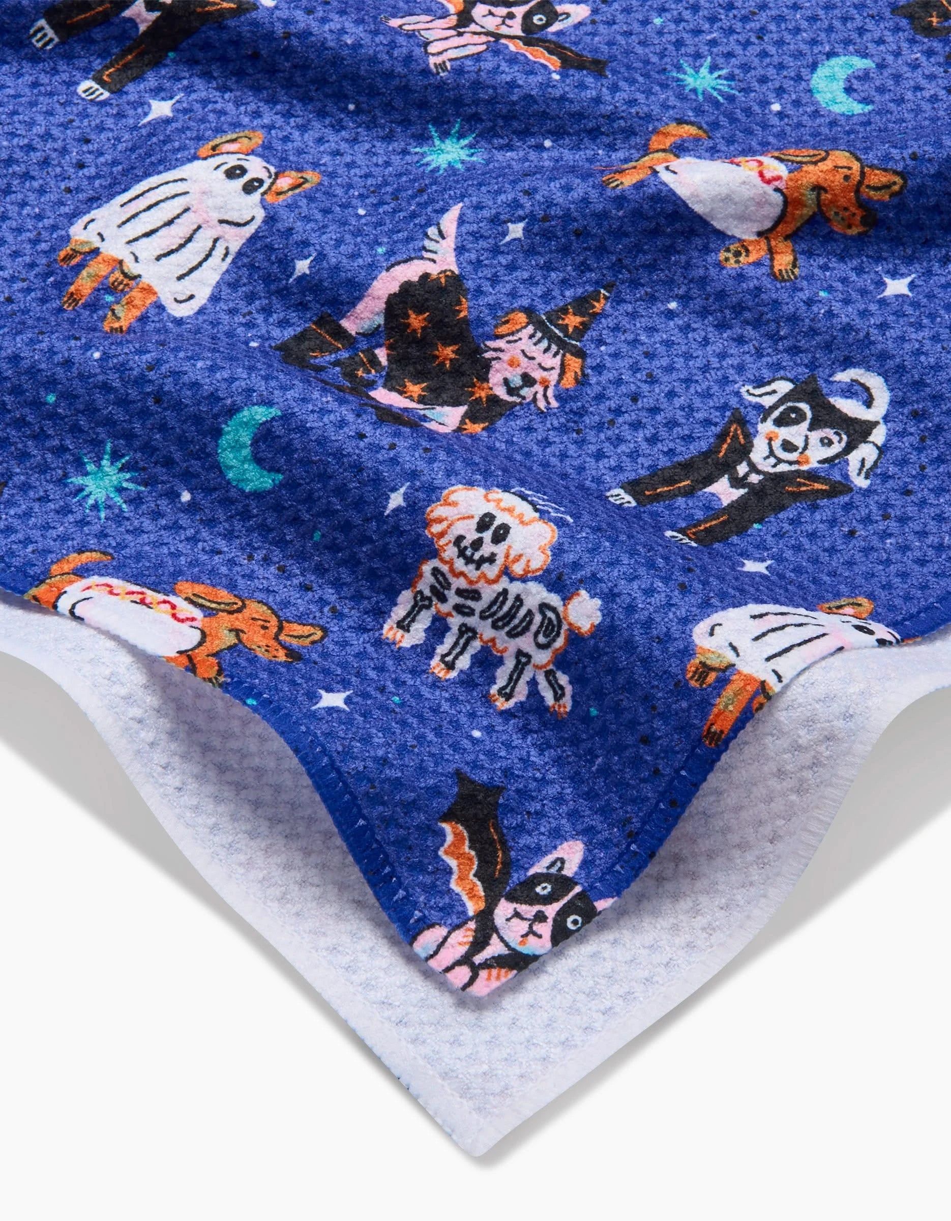 A close-up of the Camila Leão Halloween Hounds Tea Towel, made from blue Geoweave fabric and featuring cartoon dogs in Halloween costumes, stars, and crescent moons, with the white underside visible in one corner.