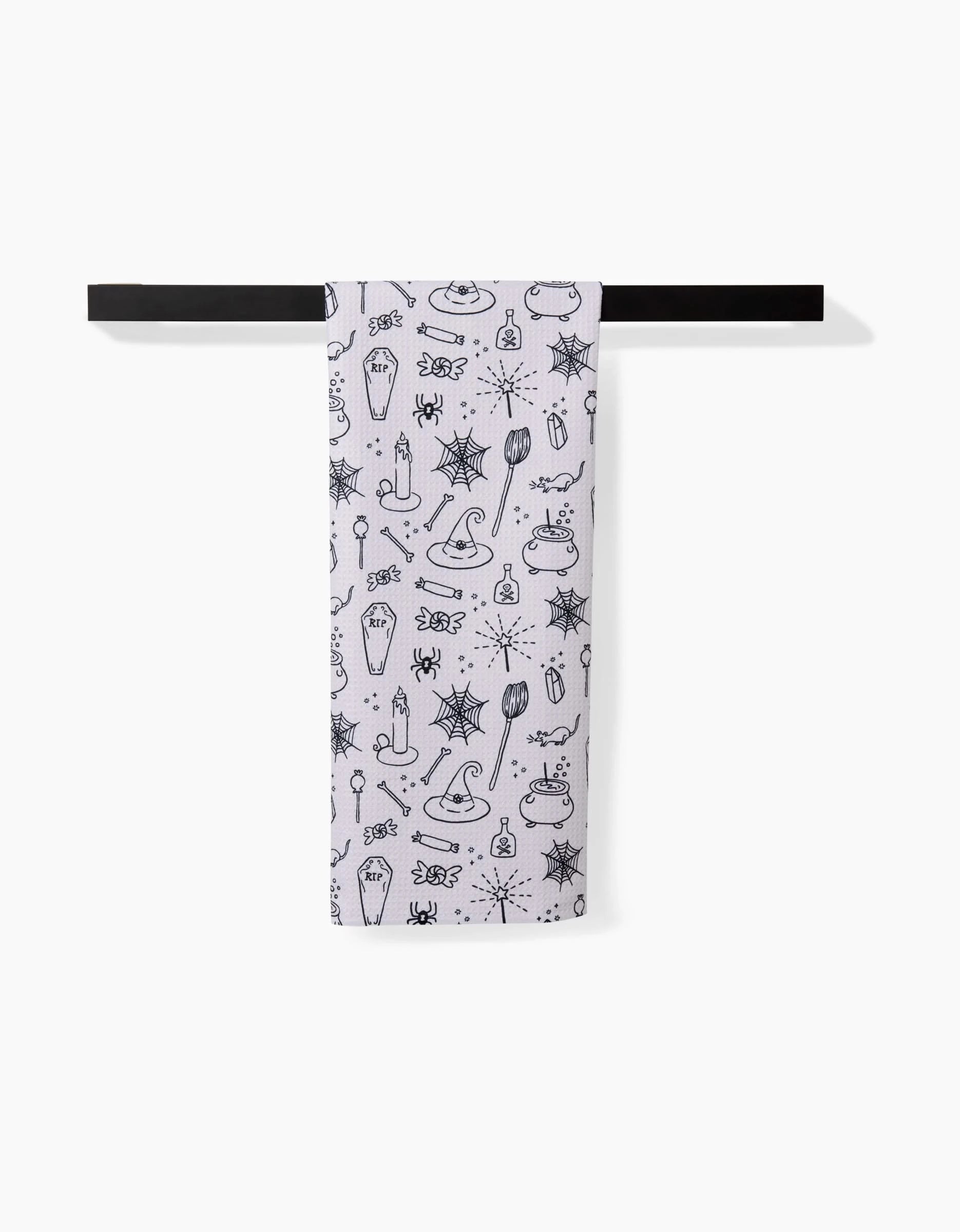 The Kayla Ann Halloween Magic White tea towel, featuring black Halloween doodles and made from recycled materials, hangs on a black rod against a plain white background.