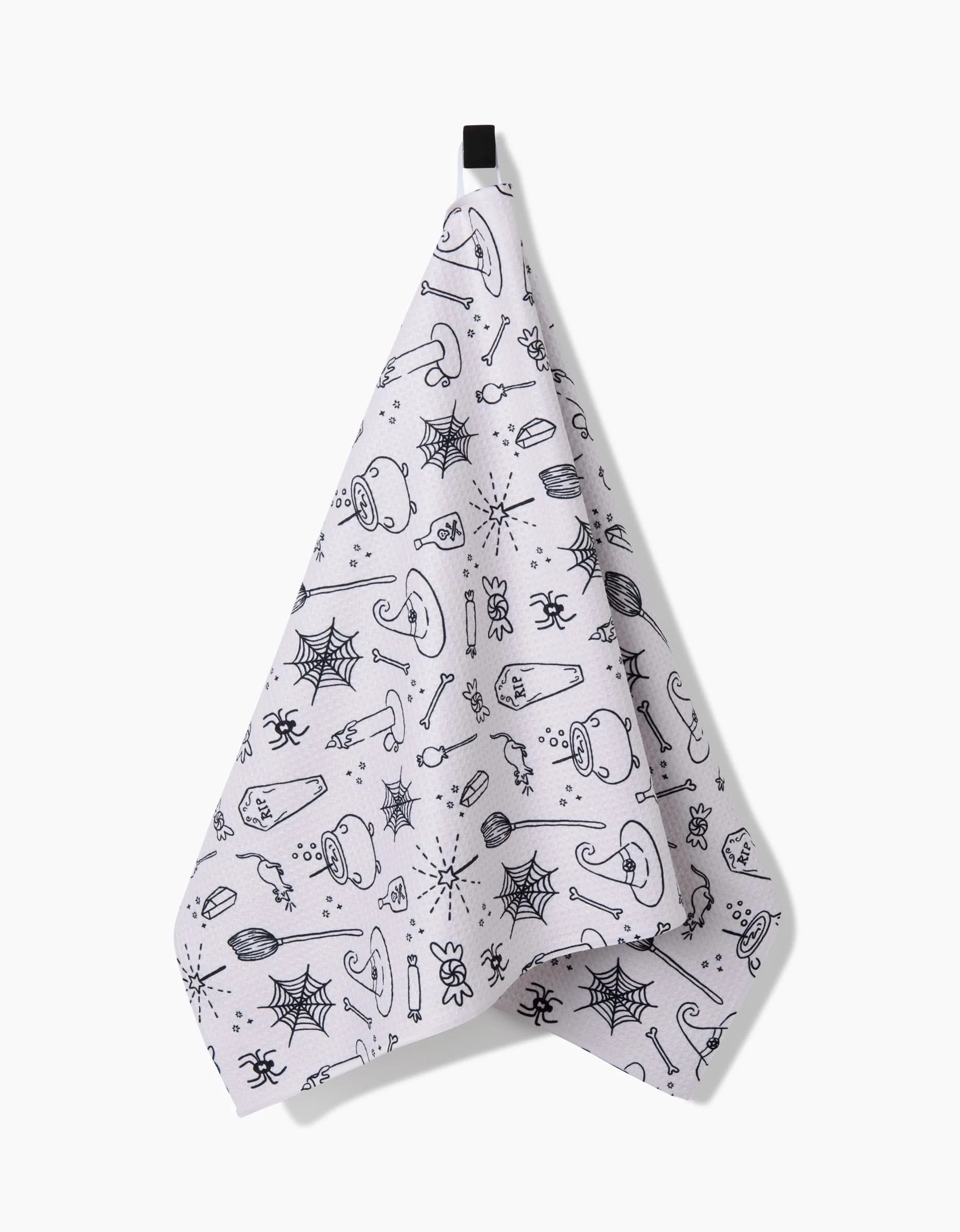 The Kayla Ann Halloween Magic White towel features ultra-absorbent Geoweave fabric with black Halloween-themed illustrations—spiders, webs, mushrooms, potion bottles, and more—hanging from a black loop on a white background.