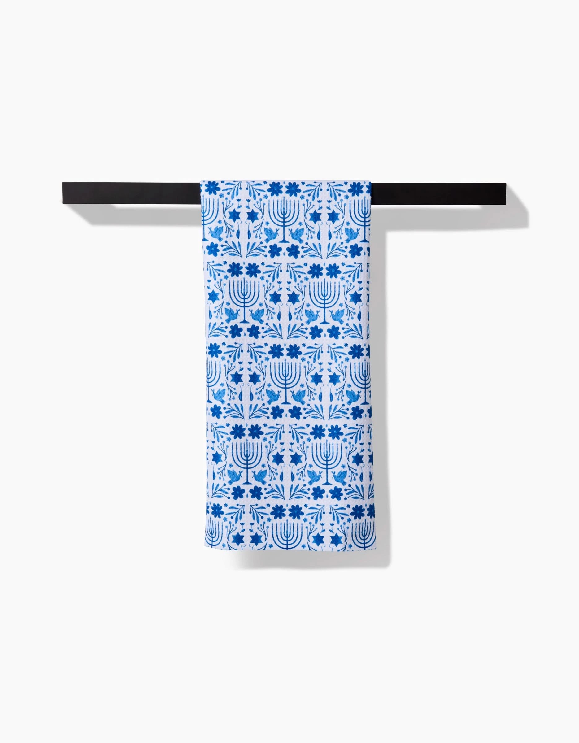 The Hanukkah tea towel by Laura Supnik, featuring a blue and white menorah and floral design made from recycled materials, is draped over a black bar against a plain white background.