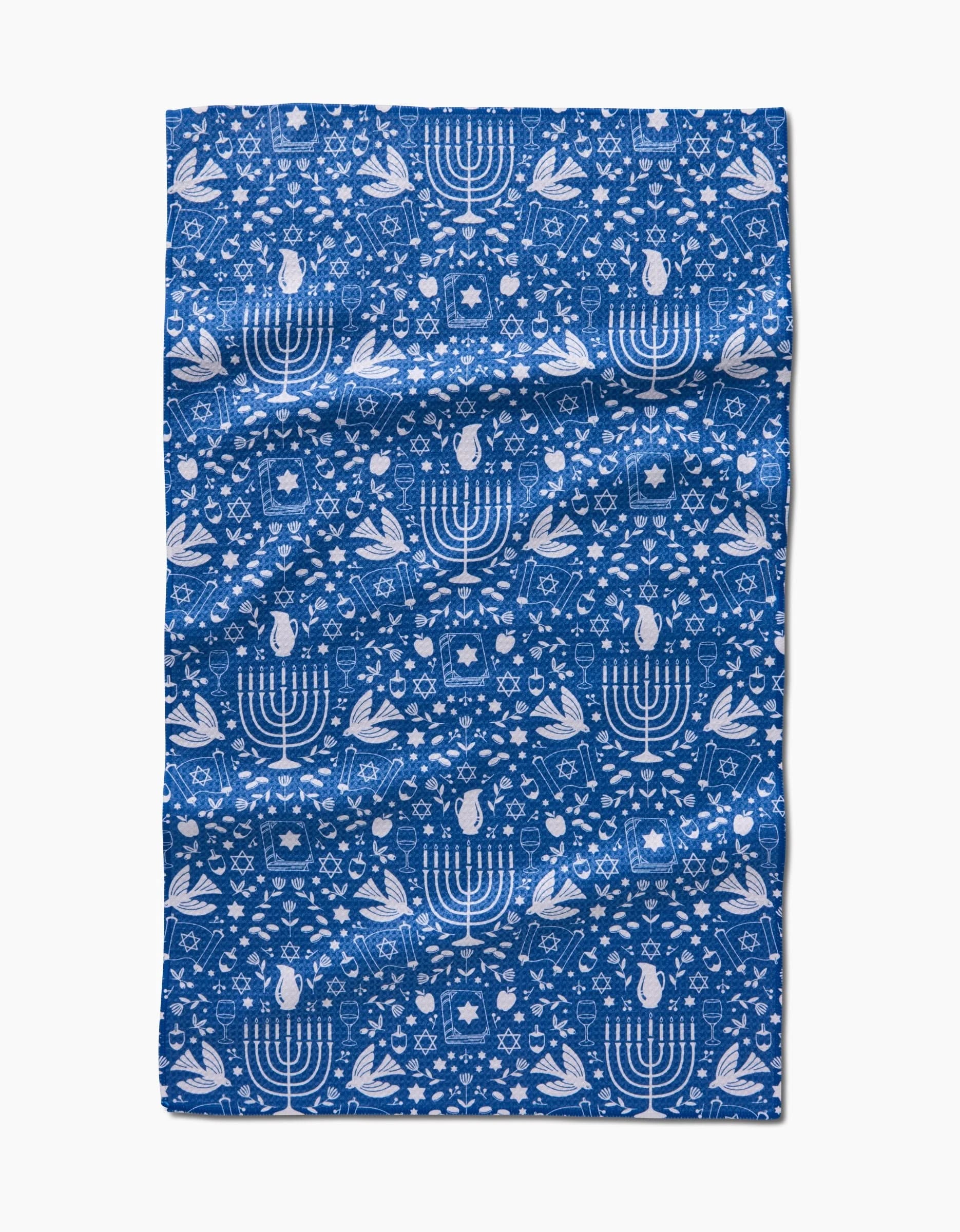 The Hanukkah Sameach kitchen towel by Megan Kampa features blue fabric with a repeating white menorah, Star of David, dove, and olive branch pattern. Made from recycled materials, it’s shown flat on a white background.