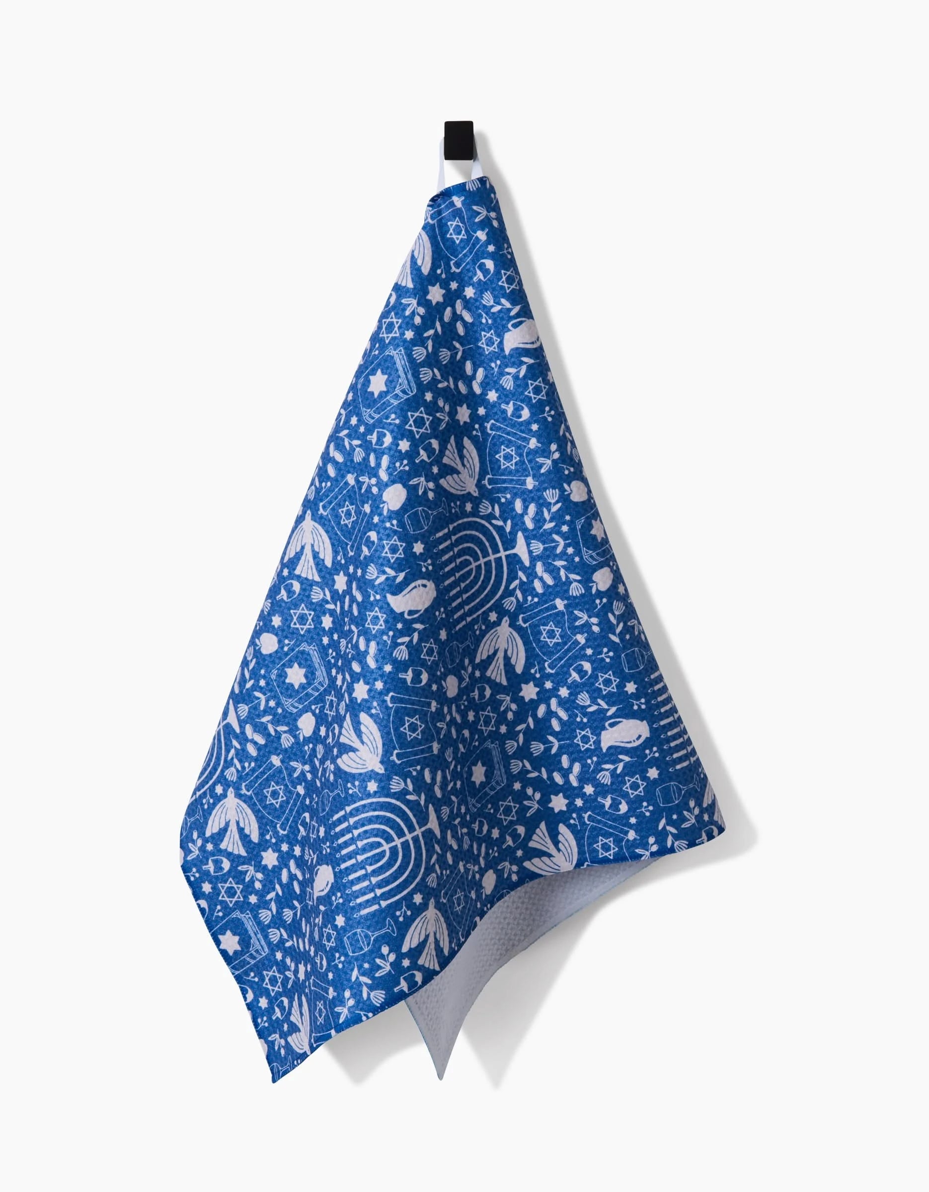 The Hanukkah Sameach tea towel by Megan Kampa features blue fabric with white illustrations of fish, birds, leaves, and geometric shapes, hanging from a black loop against a white background.