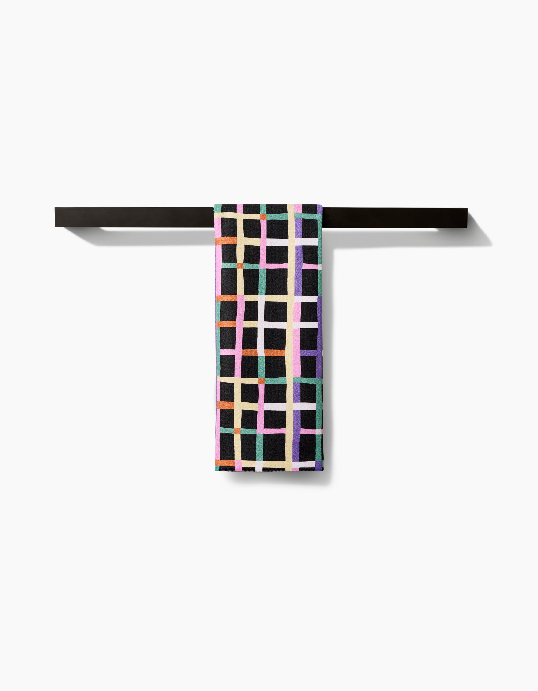 A Claire Iglesias Happy Plaid Tea Towel with a vibrant grid design hangs on a sleek black towel bar against a white background, highlighting its signature Geoweave™ fabric.