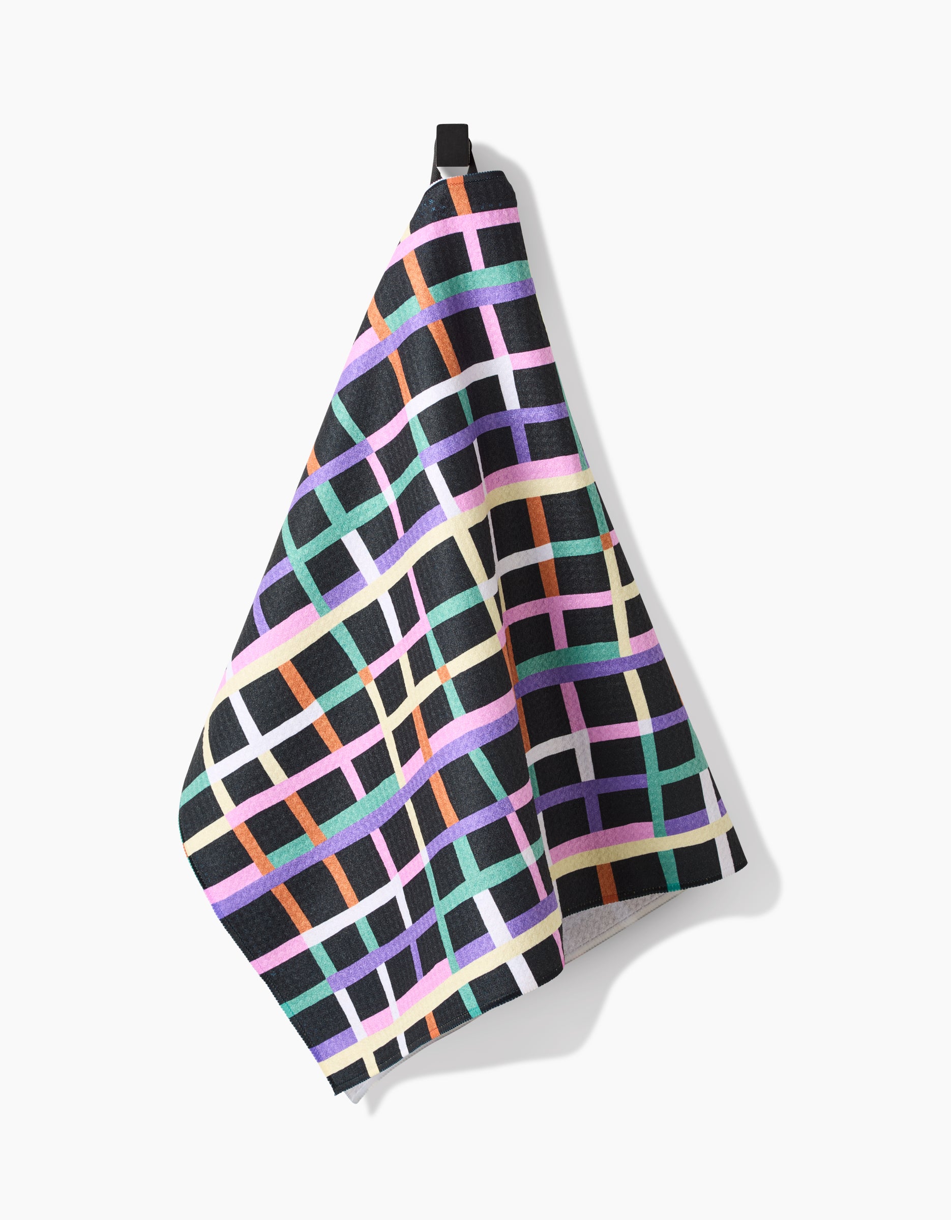 A Claire Iglesias Happy Plaid tea towel, made from Geoweave™ fabric with post-consumer recycled materials, features a colorful intersecting grid in purple, teal, orange, pink, and white. It hangs on a black loop against a white background.