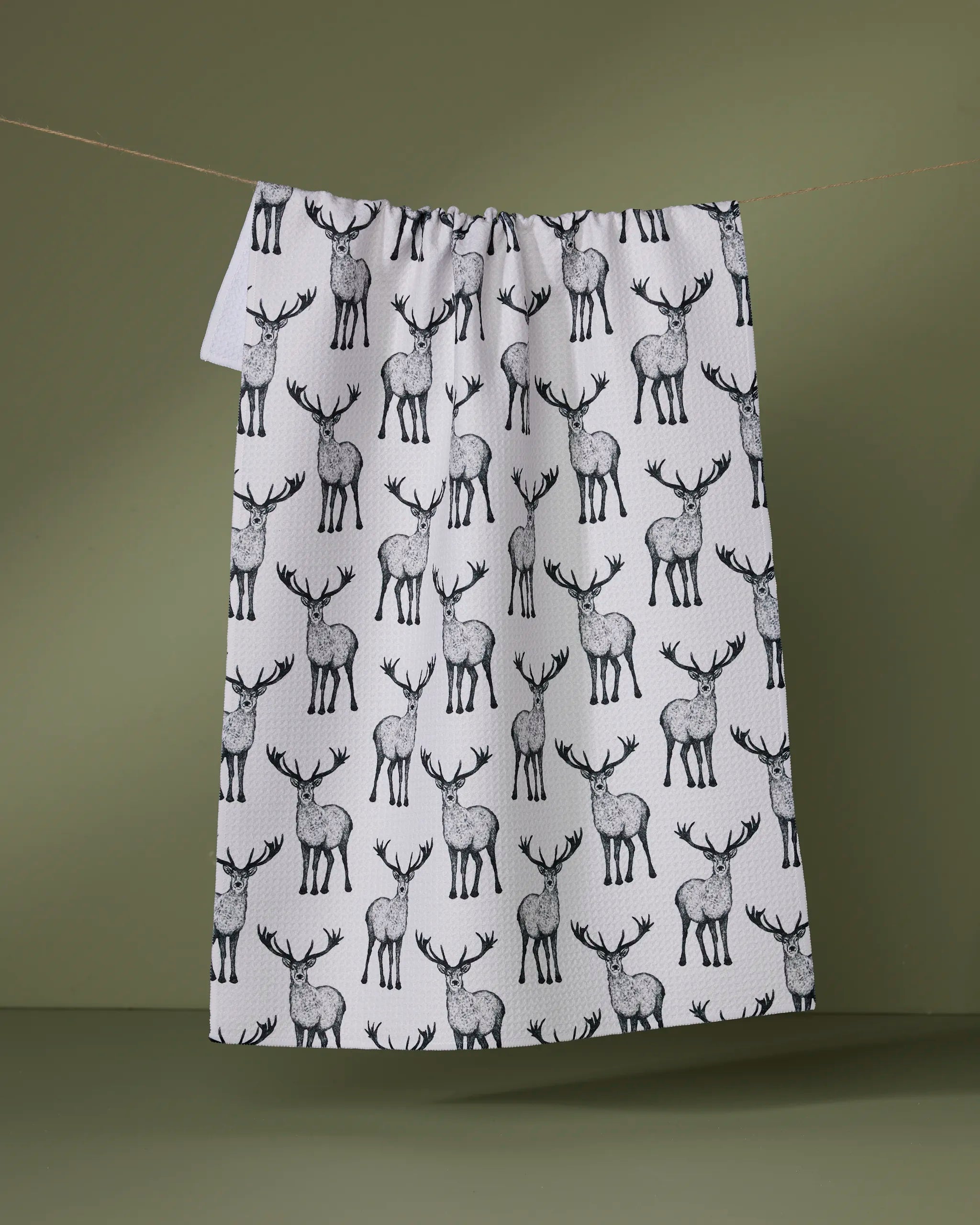 The Hartley tea towel by Geometry, made from recycled materials, displays black and grey deer in a repeating pattern and hangs against a muted green background.