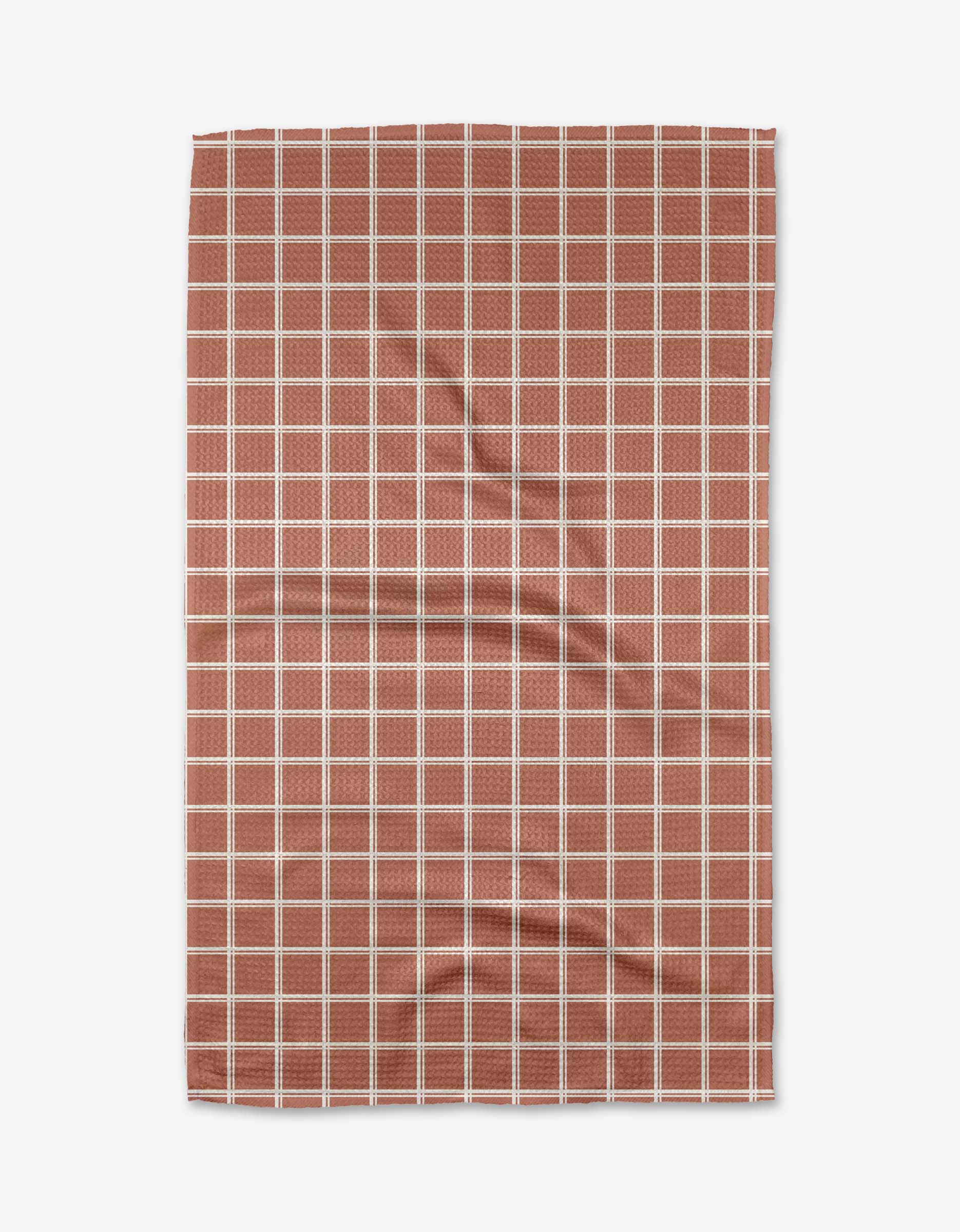 The Harvest Plaid tea towel by Charlie Rowan Designs features a reddish-brown background with an evenly spaced white grid on ultra-absorbent Geoweave fabric, shown slightly wrinkled against a white backdrop.