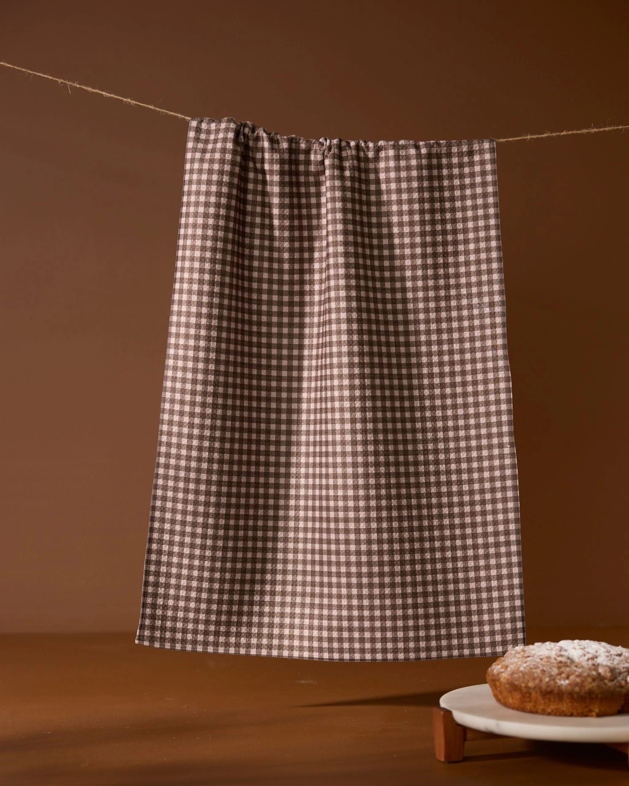 A Geometry Hearth tea towel with a brown and white checkered pattern hangs on a string against a brown background. A powdered sugar-dusted cake sits partially visible on a wooden table in the bottom right corner.