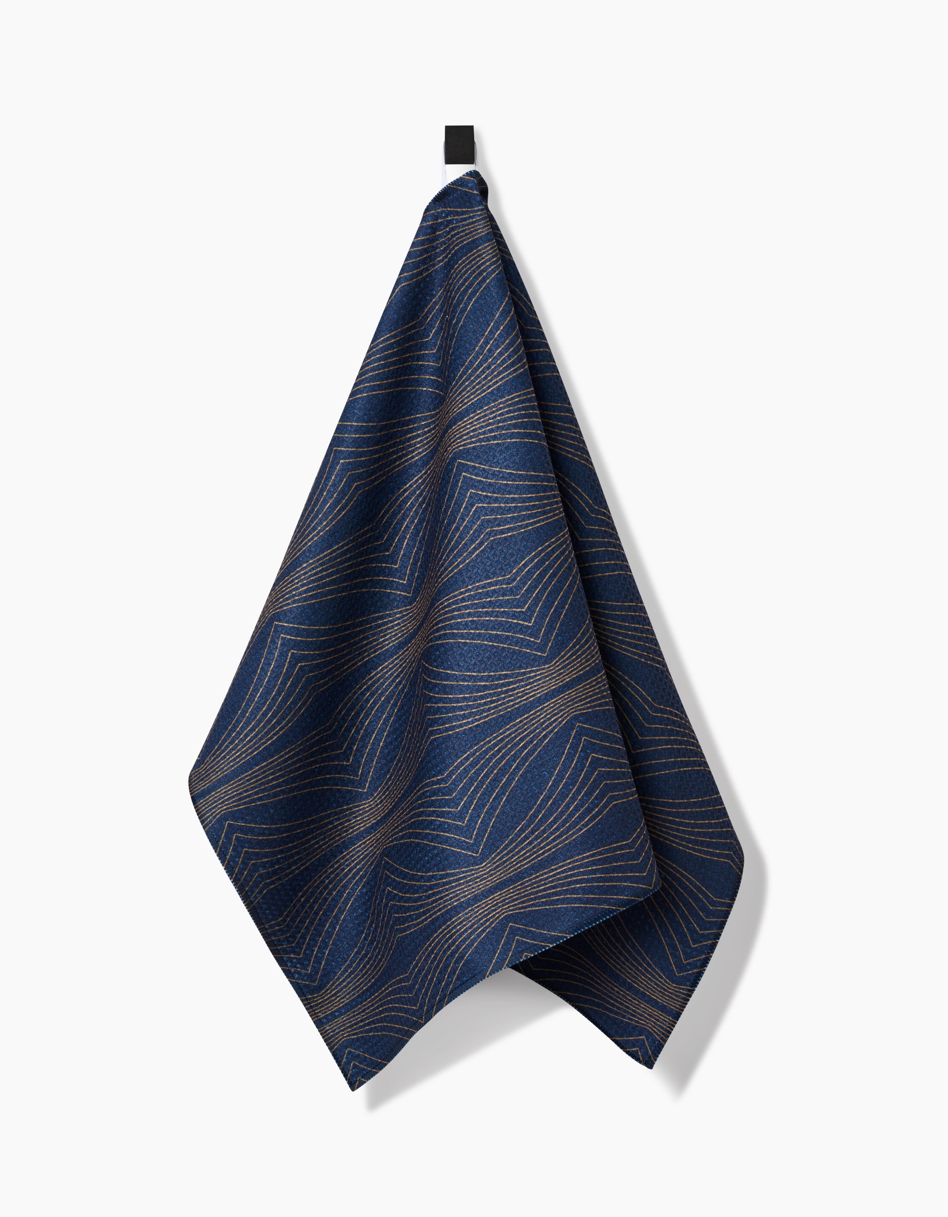 The Hera tea towel by Amy MacCready, made from Geoweave™ fabric, showcases thin tan geometric lines on dark blue and hangs by a black loop against a plain white background.