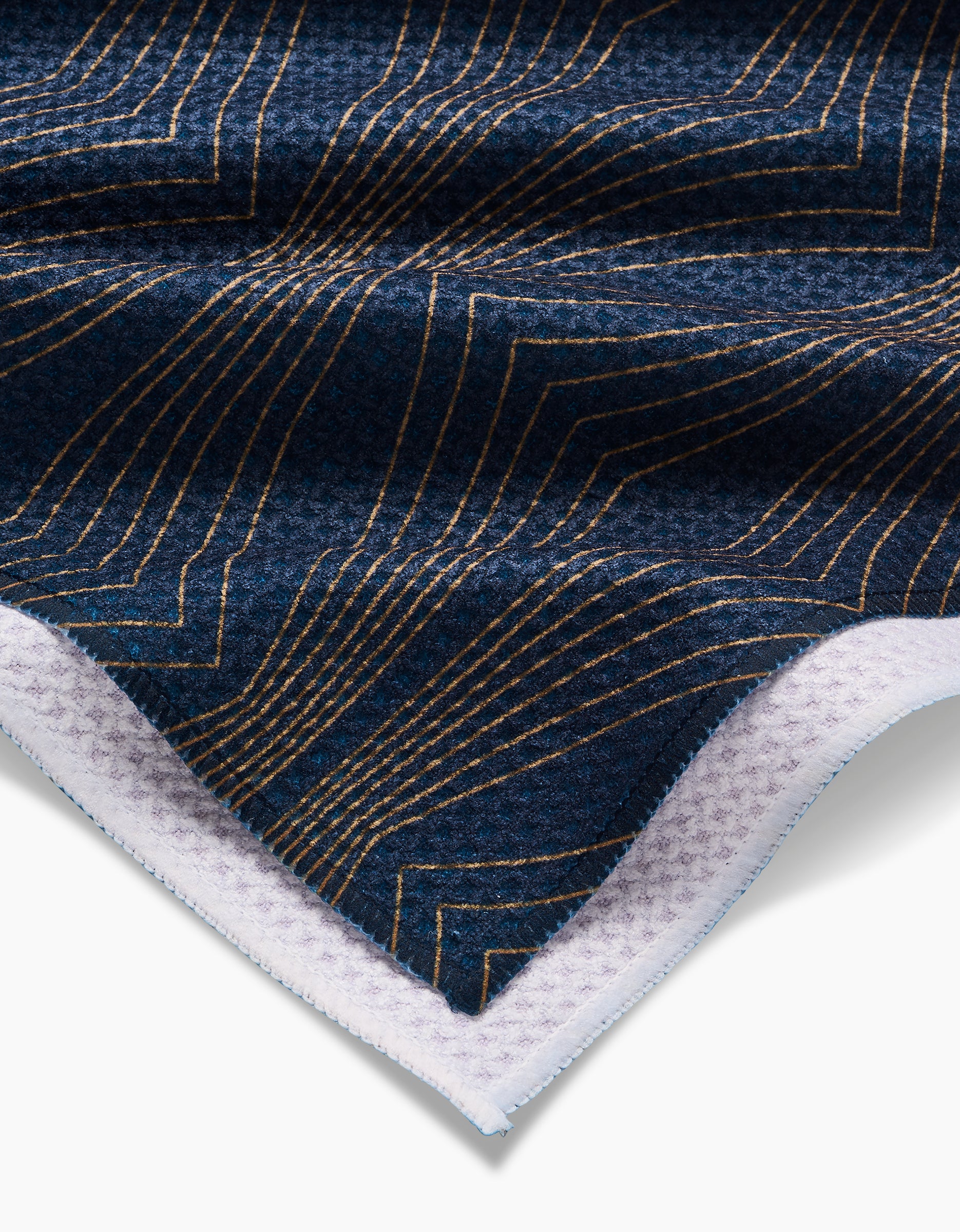 A close-up of the Hera tea towel by Amy MacCready, featuring dark blue Geoweave™ fabric with gold geometric lines and a corner folded to reveal a white, waffle-textured underside.