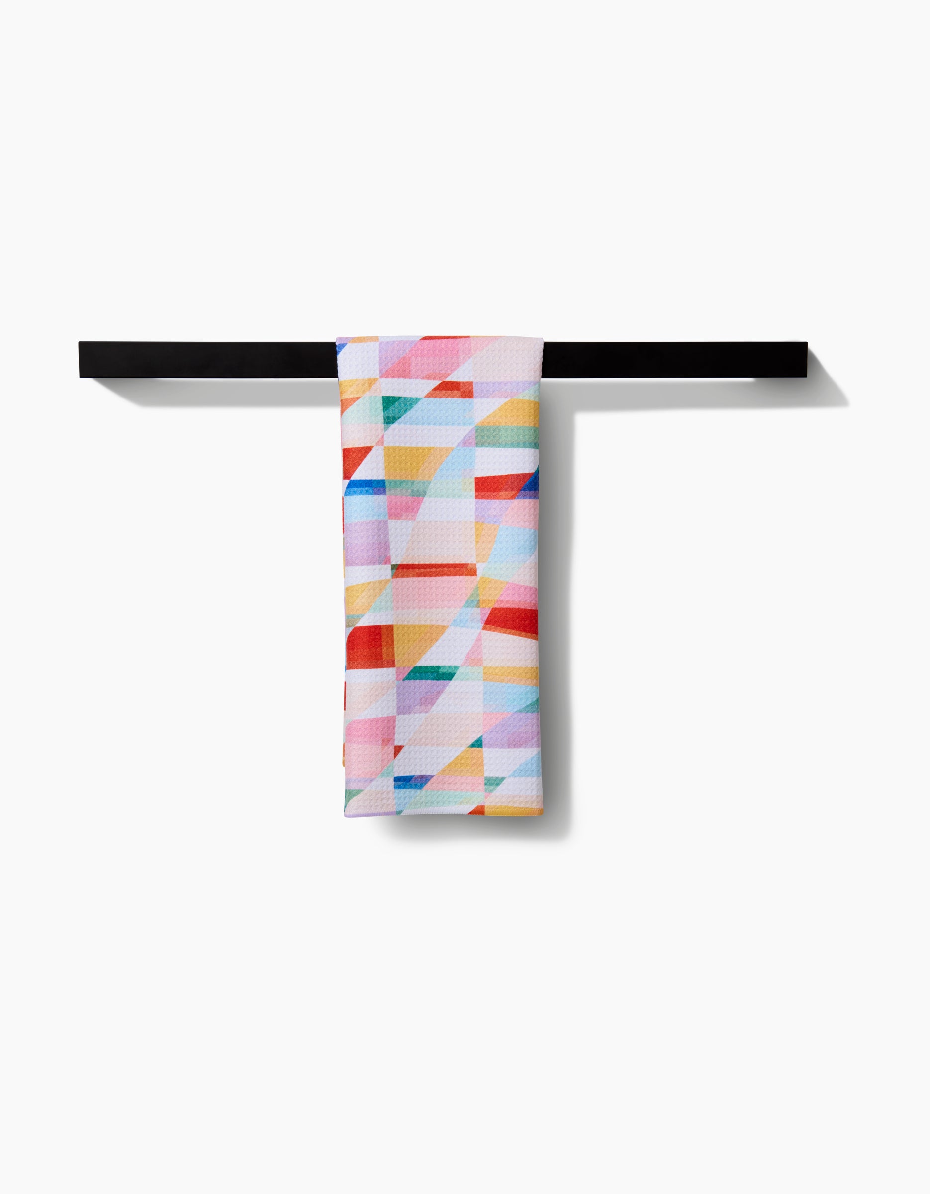 The Highlands tea towel by RACHEL PARKER, featuring a vibrant geometric pattern in red, blue, yellow, and green, hangs on a simple black bar against a white background. Made from recycled materials.