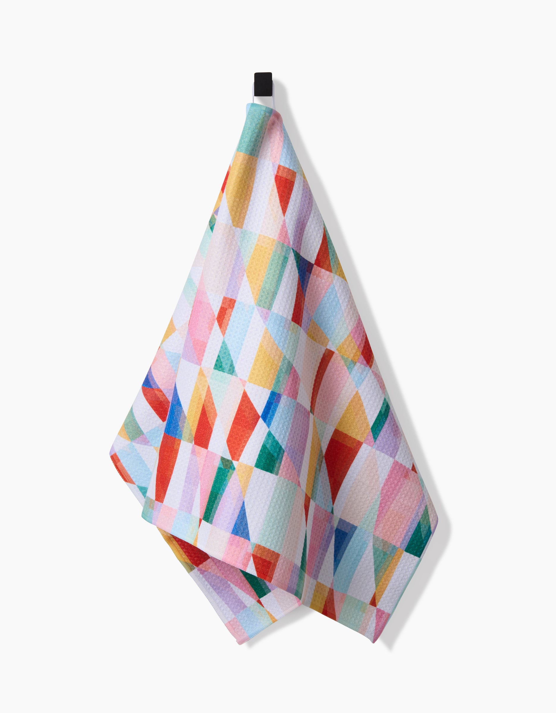 The Highlands Tea Towel by RACHEL PARKER features a vibrant geometric pattern of overlapping triangles and rectangles in bright colors, made from recycled materials, and hangs from a black loop against a white background.