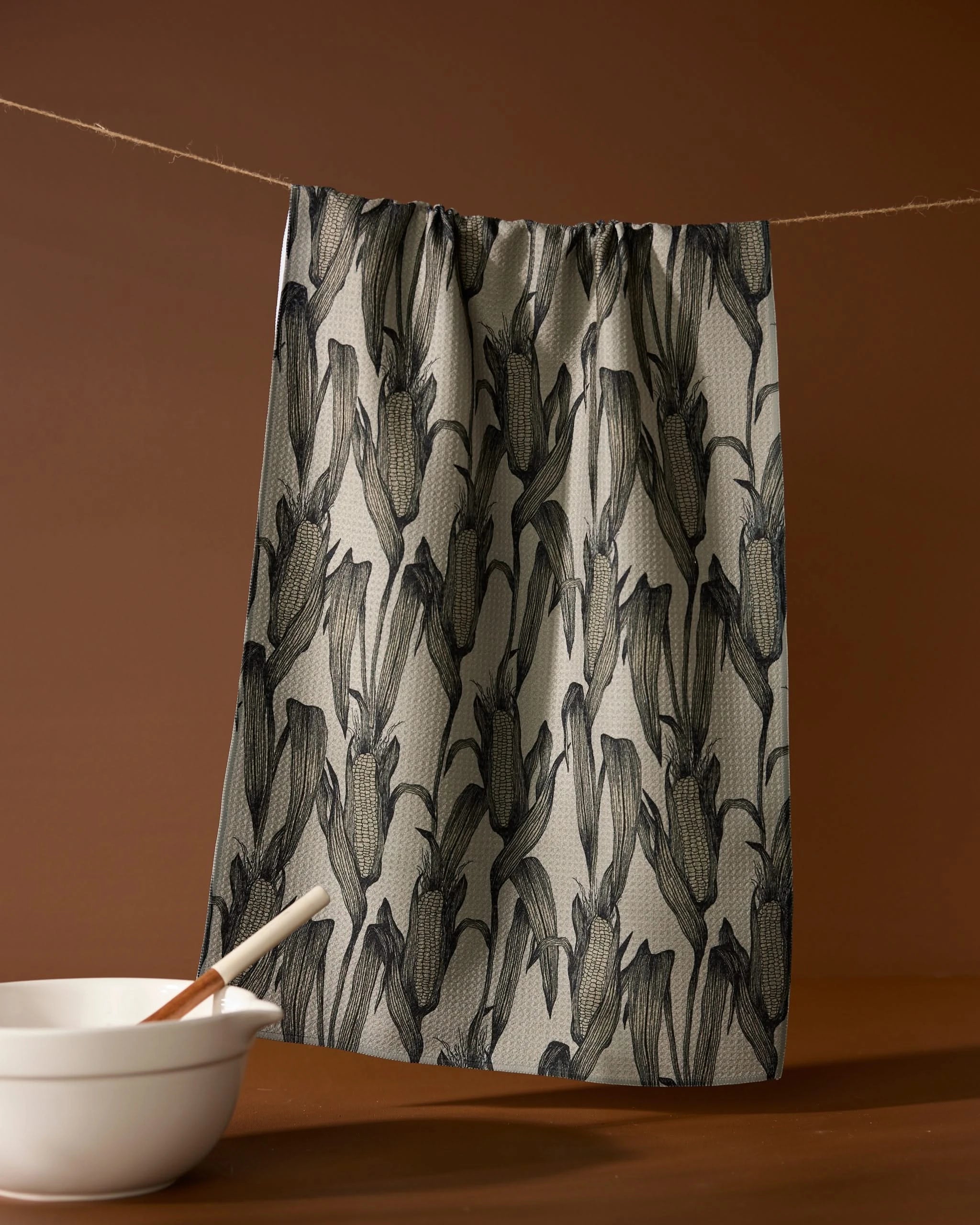 The Geometry Husk recycled tea towel, featuring a black botanical pattern, hangs on a string against a brown backdrop. A white bowl with a wooden spoon sits in front, enhancing the inviting kitchen scene.