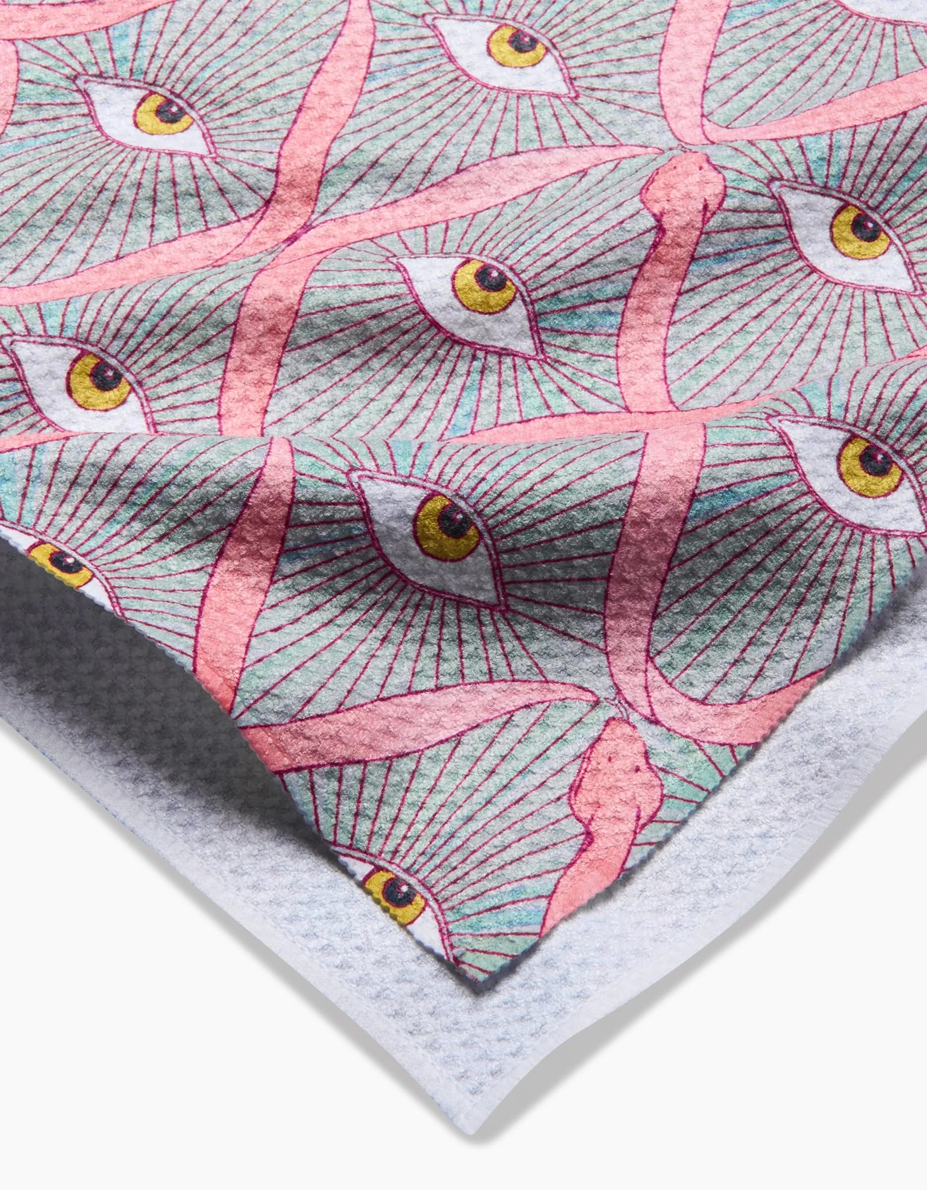 A close-up of the Illusionist Geoweave tea towel by Maayan Alper-Swan showcases yellow eyes, pink lines, and snake-like shapes on blue. A folded corner highlights its unique pattern and sustainable recycled materials.