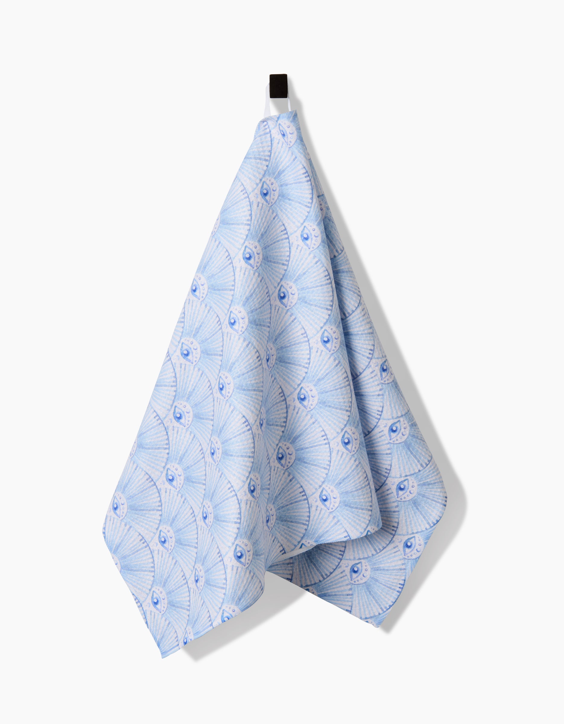 A blue and white patterned Into The Mystic tea towel by Maayan Alper-Swan, made from Geoweave fabric with a seashell-inspired design, hangs from a black hook against a plain white background.