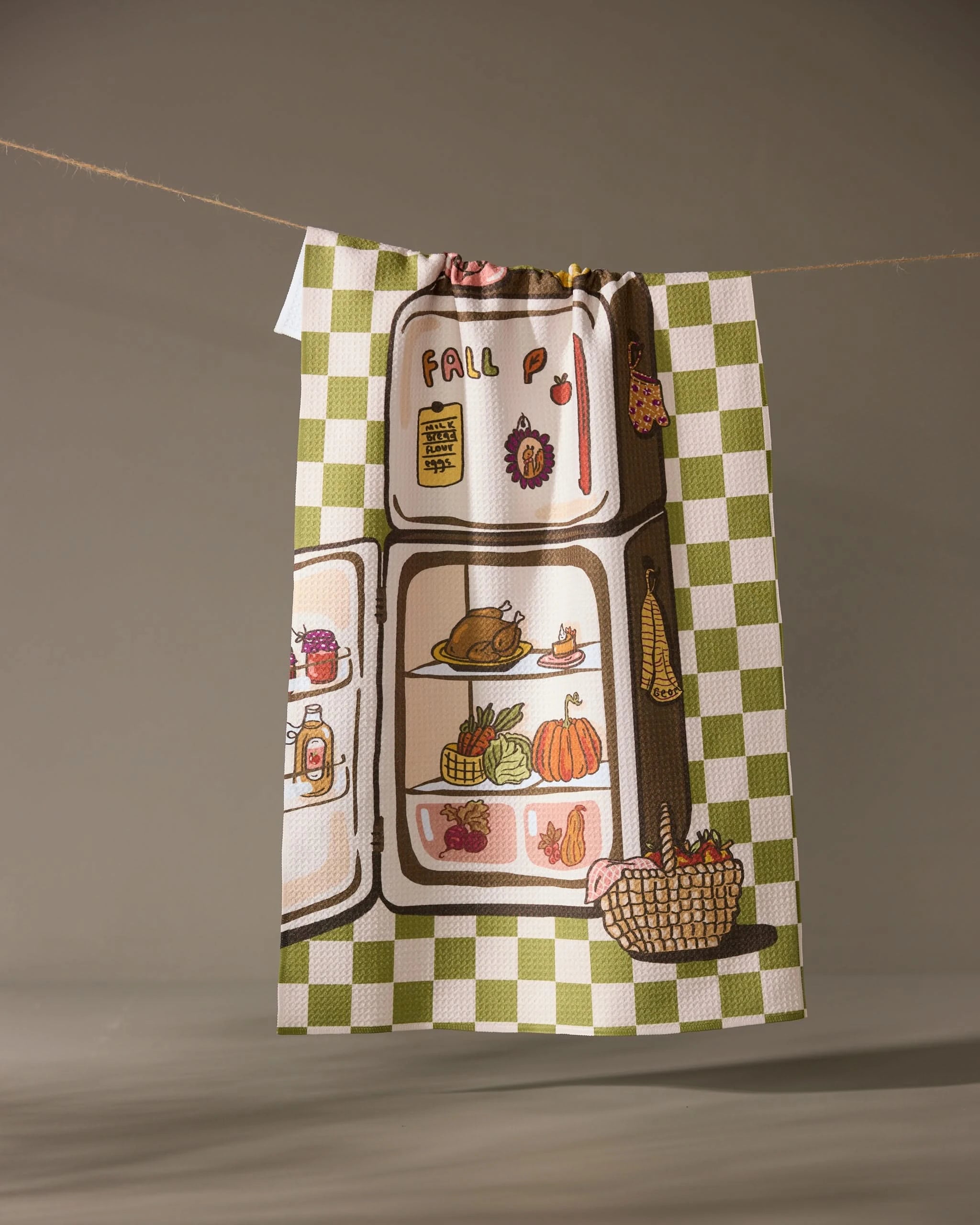 The Geometry Is It Fall Yet tea towel features a green and white checkered border and a refrigerator filled with fall foods like turkey, pumpkin, and corn, hanging on a clothesline.