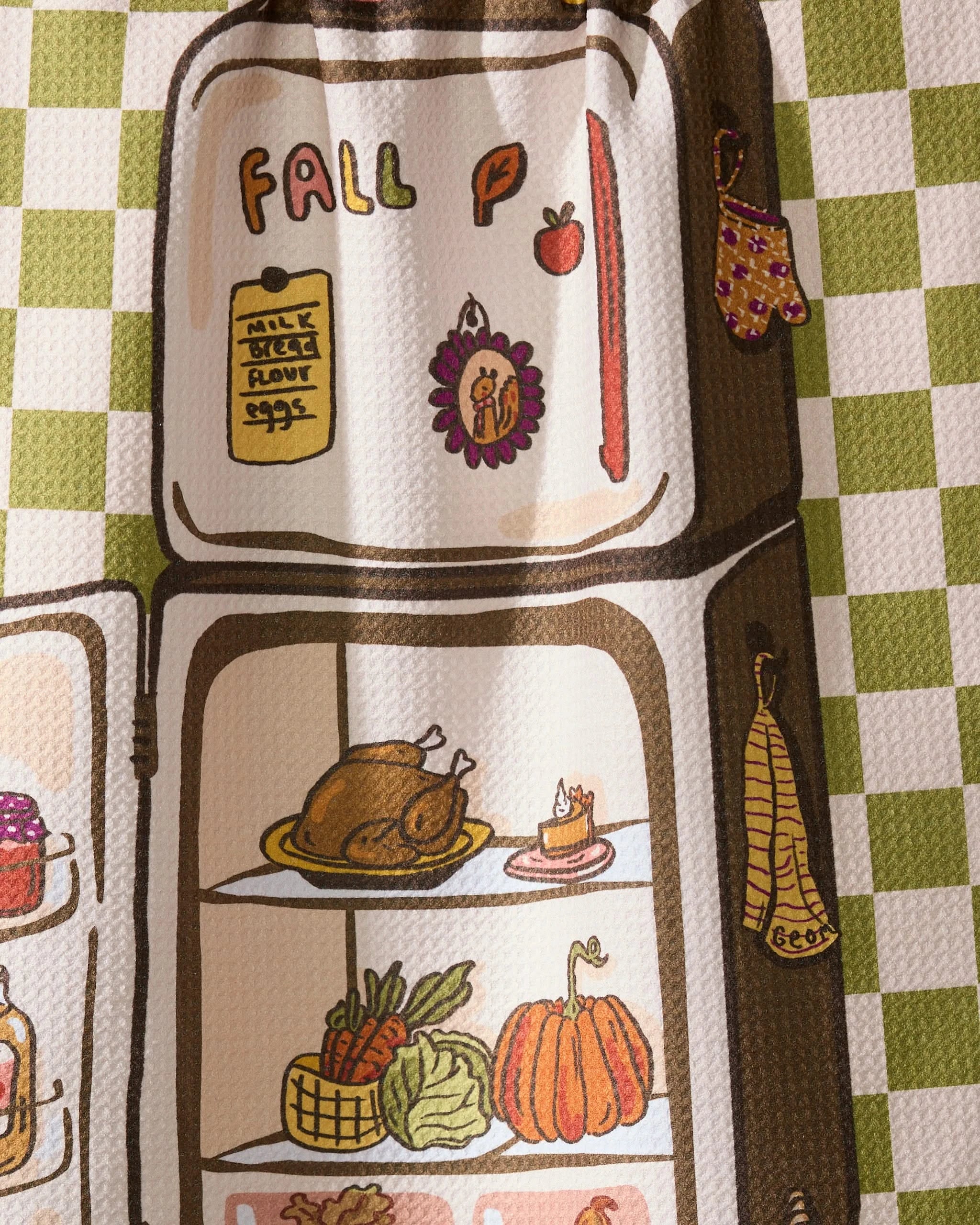 Geometry’s “Is It Fall Yet” tea towel features a vintage fridge on a green checkered background, stocked with roast turkey, pie, pumpkin, bananas, and veggies. Fall icons and an oven glove on the door create cozy autumn kitchen vibes.