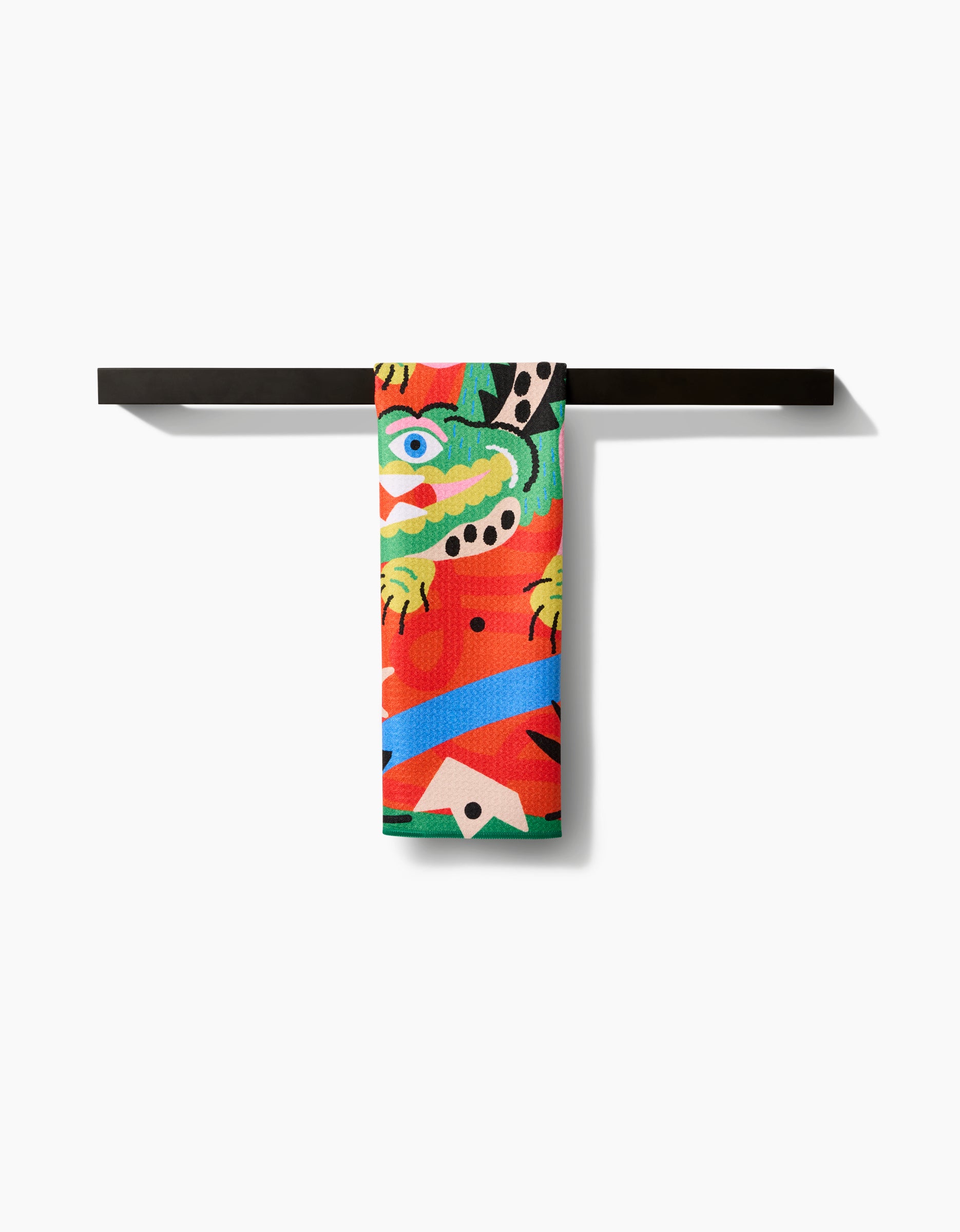 The Jacaré Tea Towel by Camila Leão, crafted from Geoweave fabric with recycled materials, features abstract patterns and an eye. It hangs neatly over a minimal black towel bar against a white background.