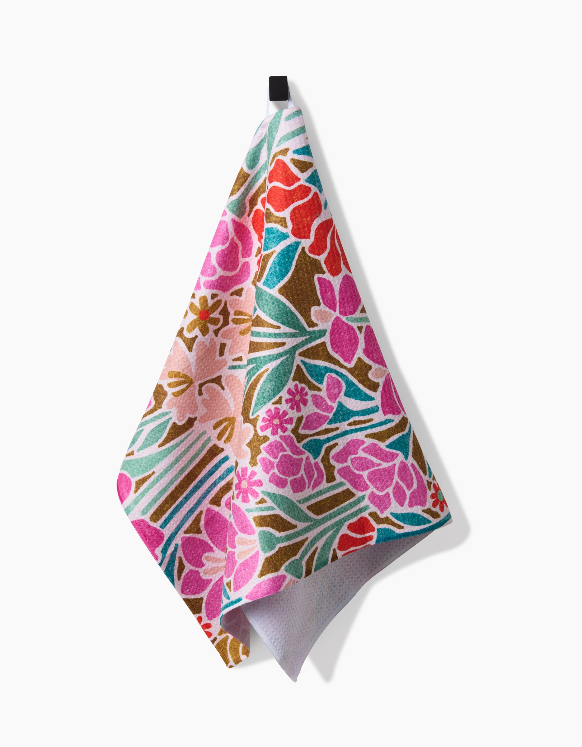 The Janae tea towel by Amy MacCready features a vibrant floral pattern in pink, red, green, and gold. Crafted from recycled materials, it hangs from a black hook against a white background.