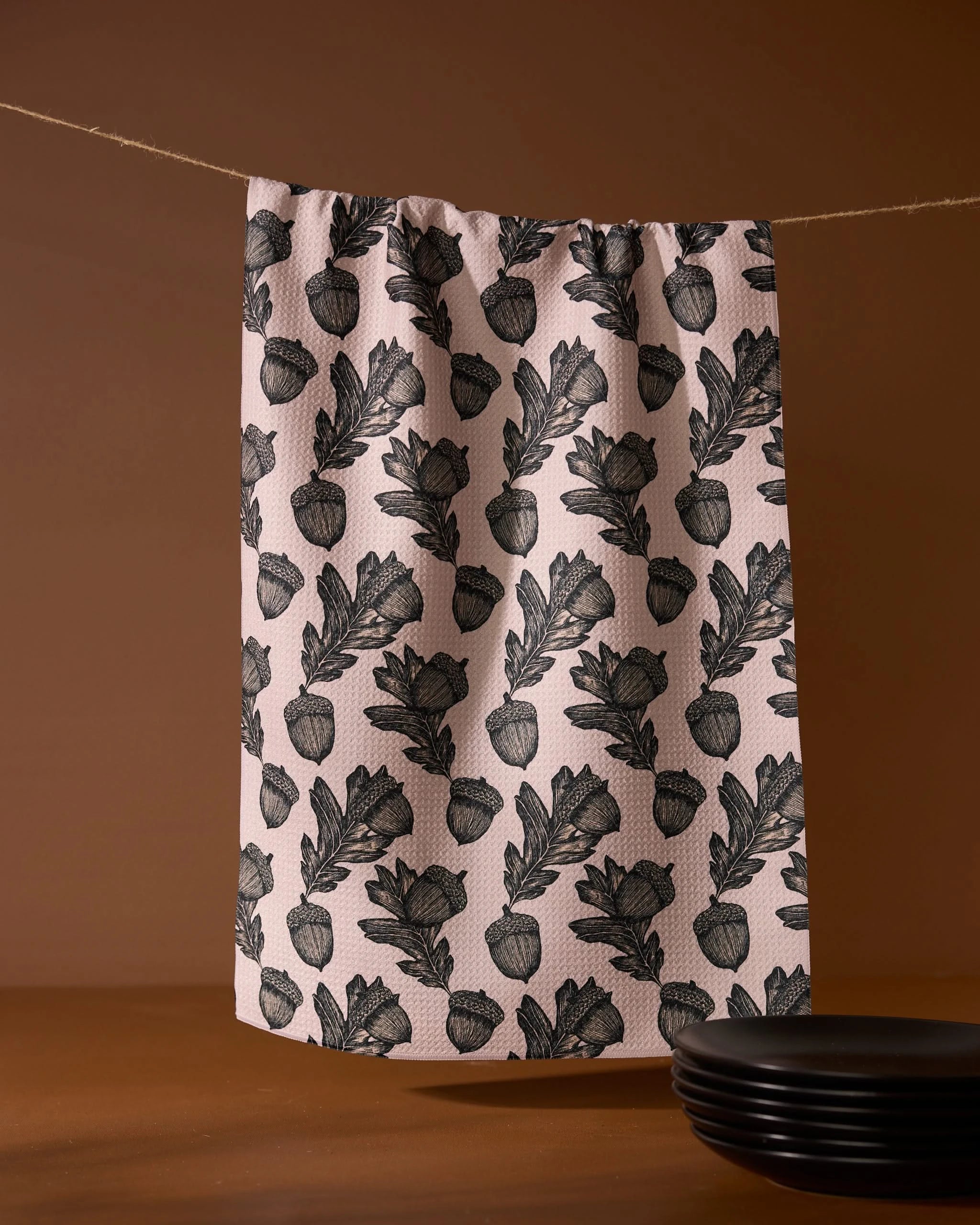 The Kindred kitchen towel by Geometry, featuring a black botanical pattern on pink recycled fabric, hangs on a clothesline against a brown background, with black plates stacked in the lower right corner.