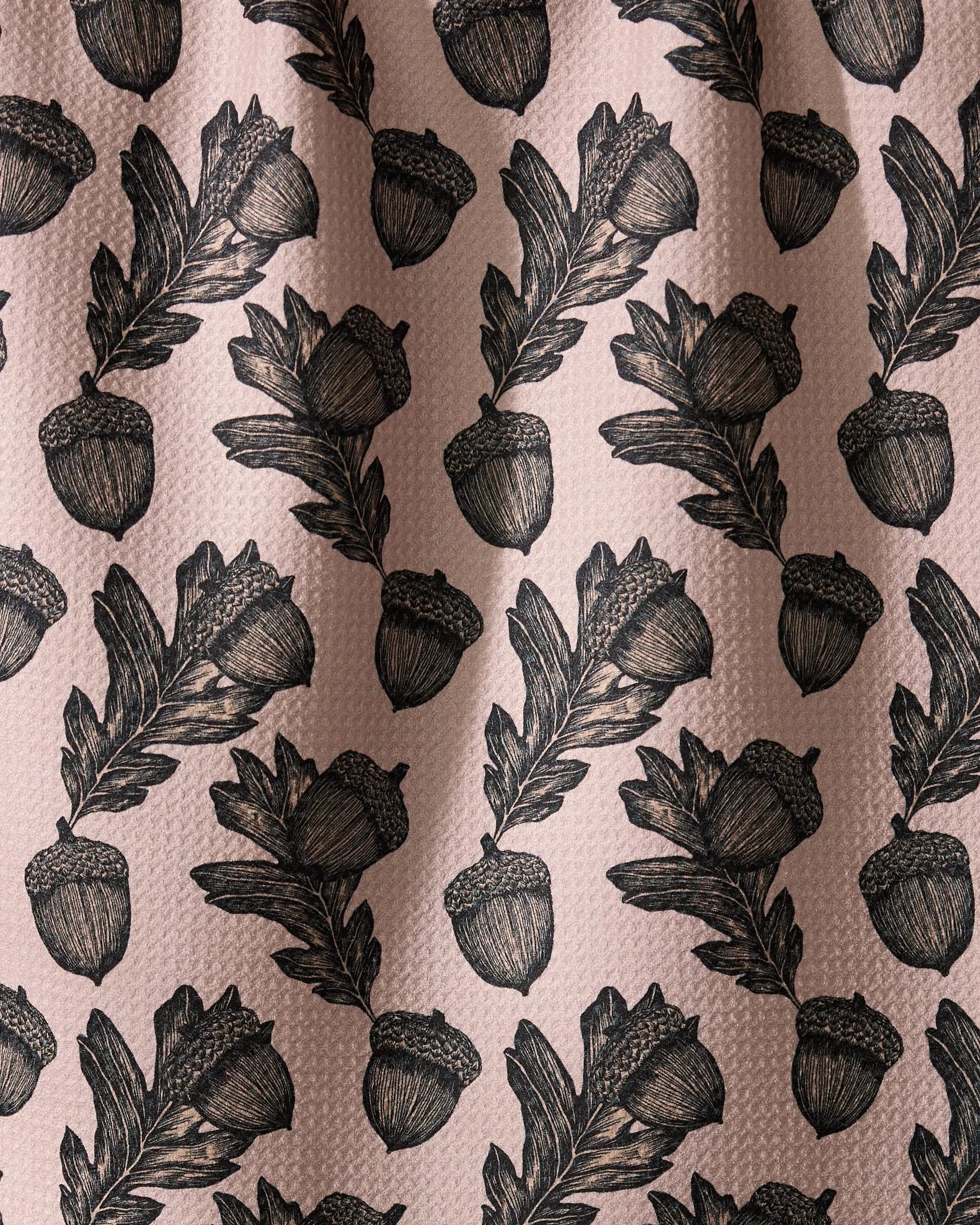 Geometry’s Kindred dish towel features an illustrated pattern of black acorns and oak leaves, evenly spaced on a textured light pink background. Ultra-absorbent fabric forms soft folds with subtle shadows and highlights.