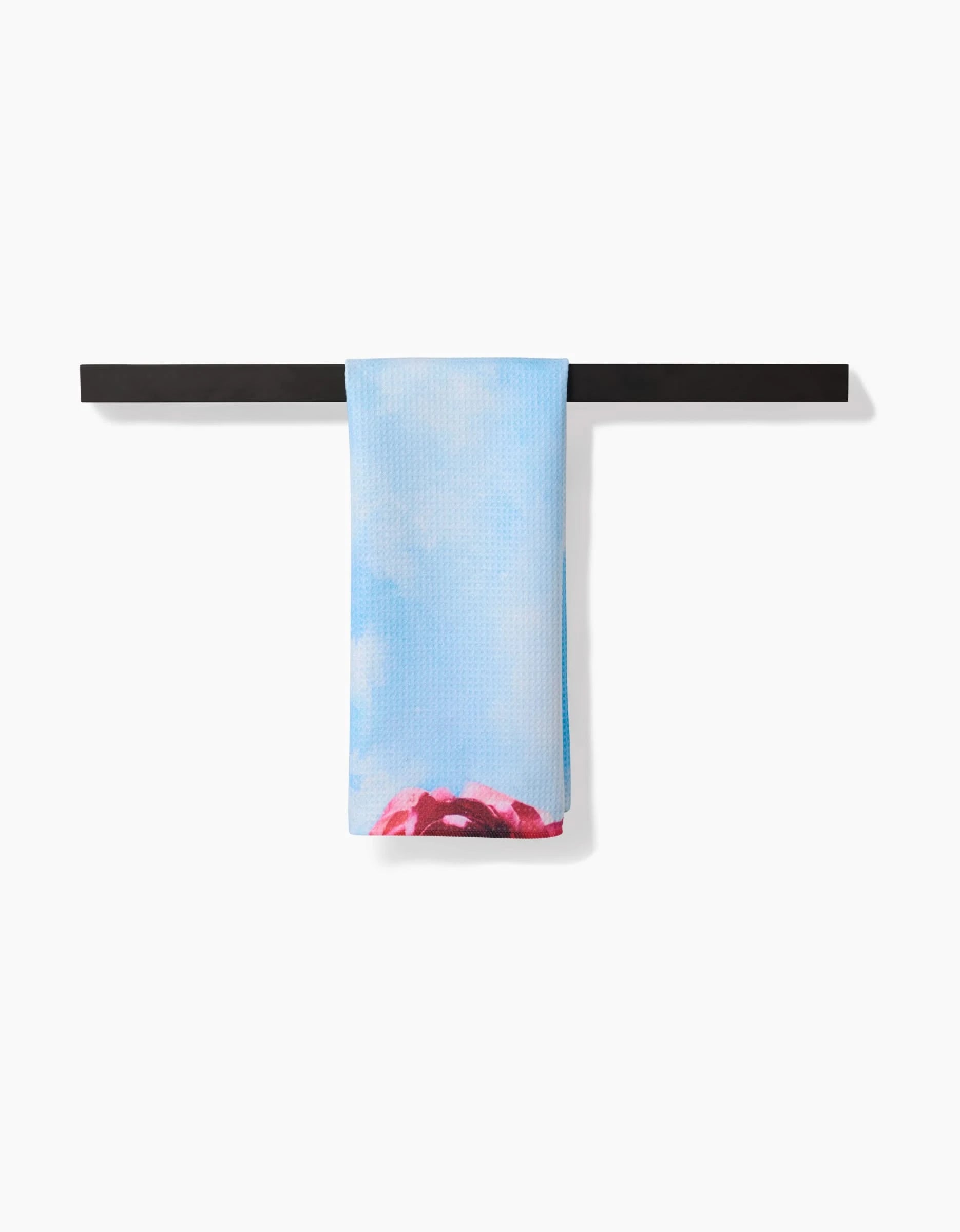 The La Floraison tea towel by Stefanie Bales, crafted with Geoweave™ fabric in light blue with a sky-and-cloud design and red accent, hangs neatly on a black bar against a white background.