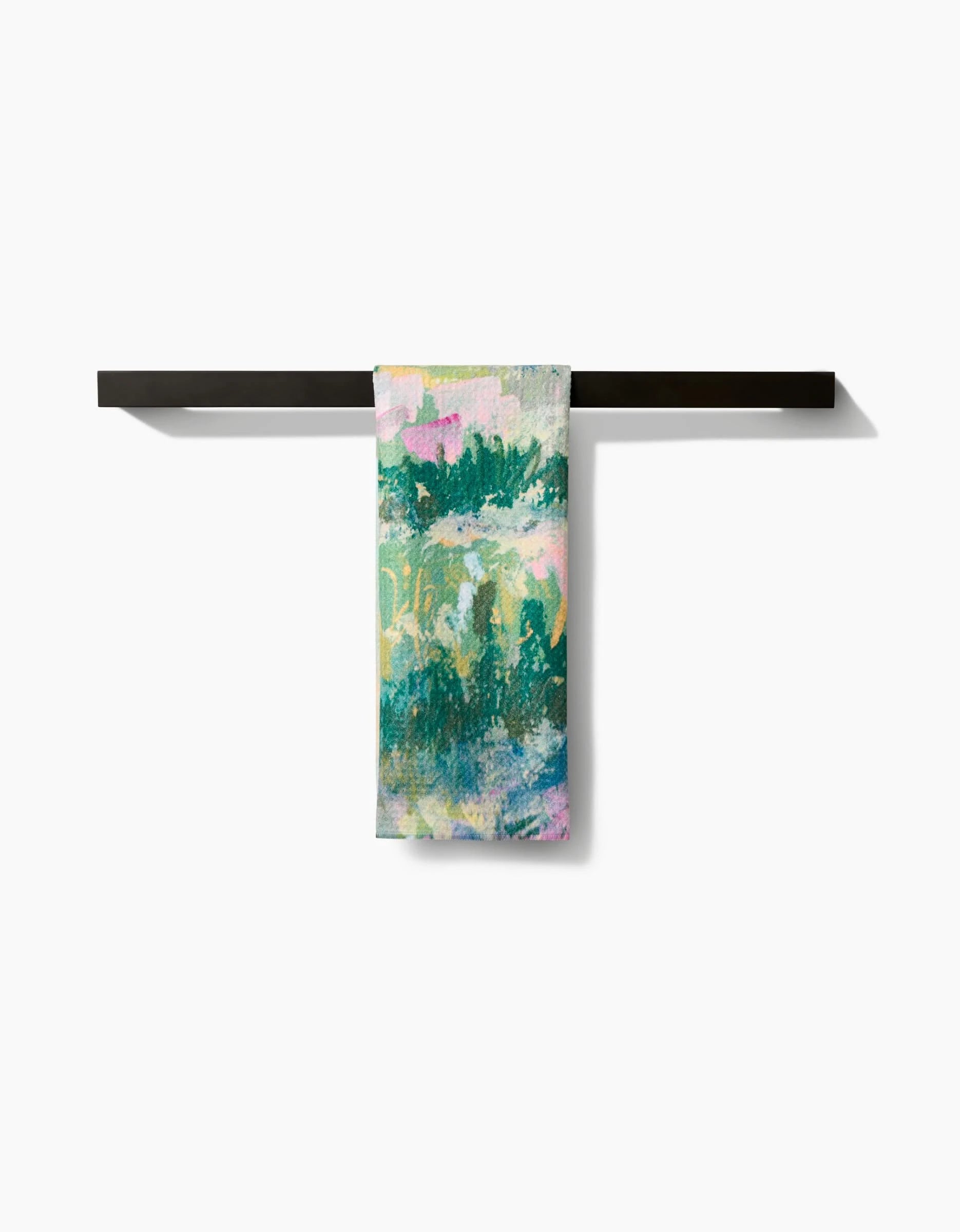 The Land of Milk and Honey Kitchen Tea Towel by Taryn Brennan, featuring green, yellow, white, and pink abstract patterns, hangs neatly on a black towel bar. Made from recycled materials and shown against a plain white background.