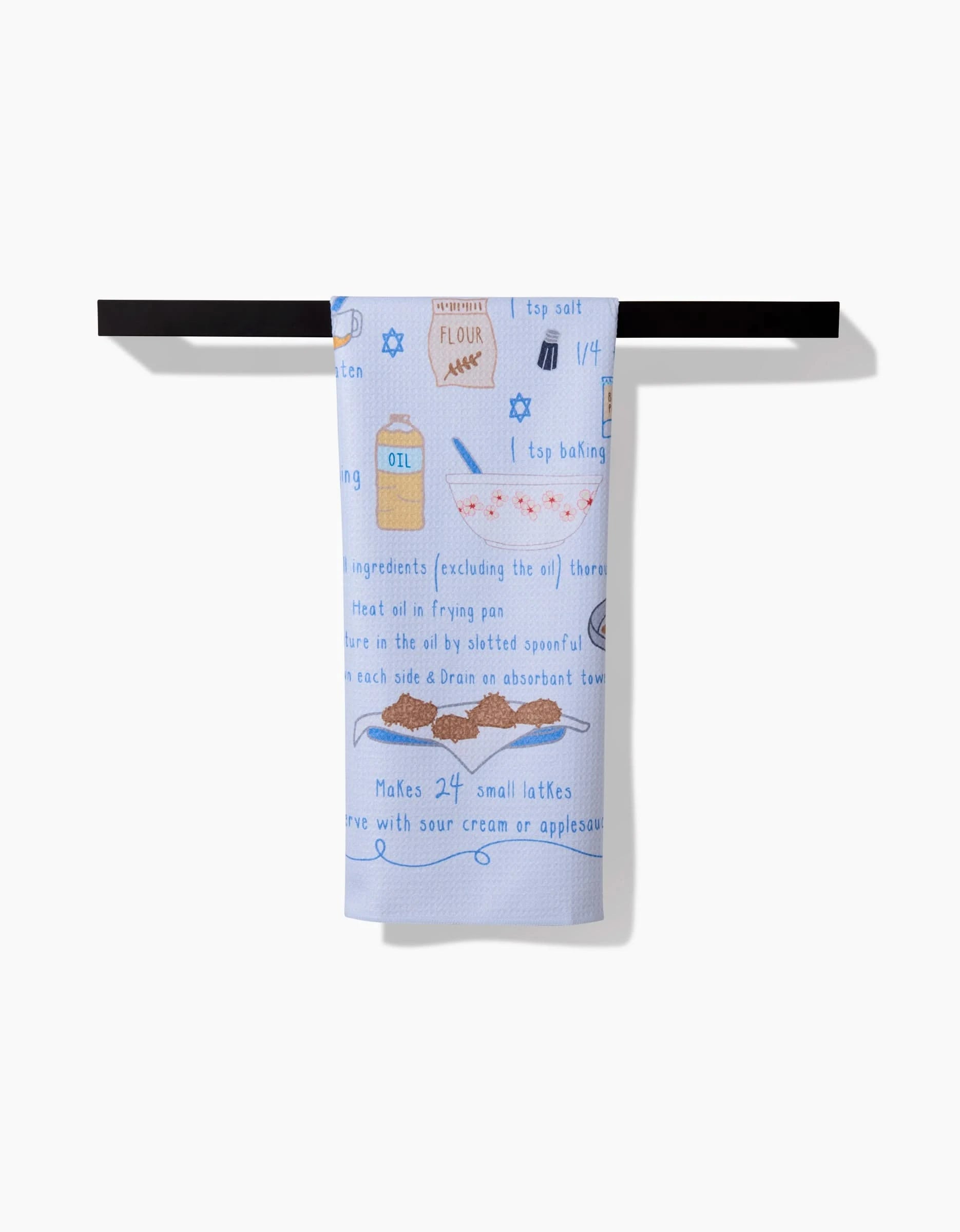 The Mindfulnice Latke Love kitchen towel, made from recycled materials and featuring illustrated instructions and ingredients—flour, oil, salt, eggs—hangs on a horizontal black rod against a white background.