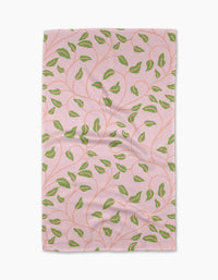 The Leafy Maze kitchen towel by Jasmine Blooms Designs is a rectangular pink recycled towel featuring green leaf patterns and winding orange stems, shown laid flat with some visible creases.