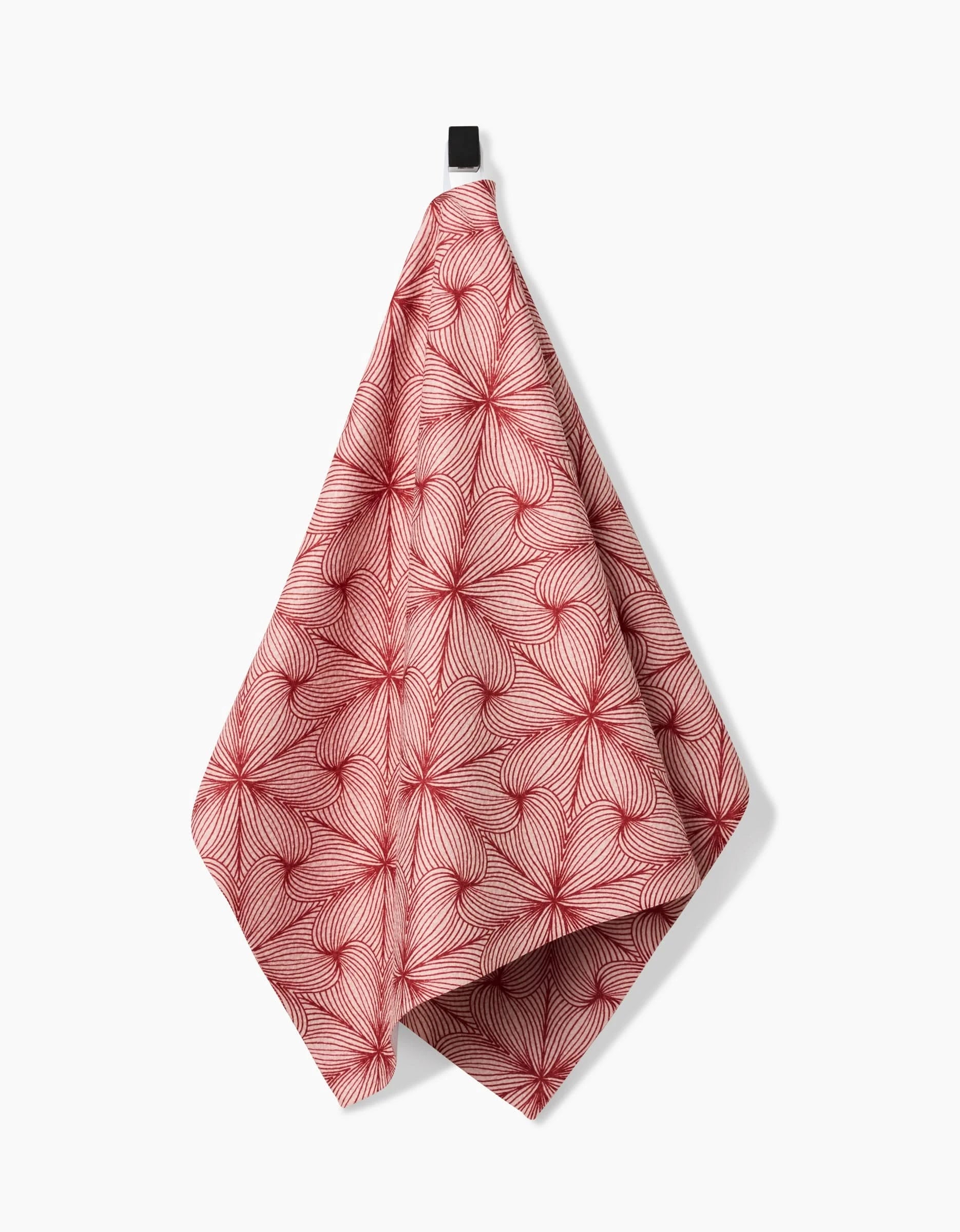 The “Lined” tea towel by Marta Harvey features a red and white geometric swirl pattern and hangs from a black loop against a white background.