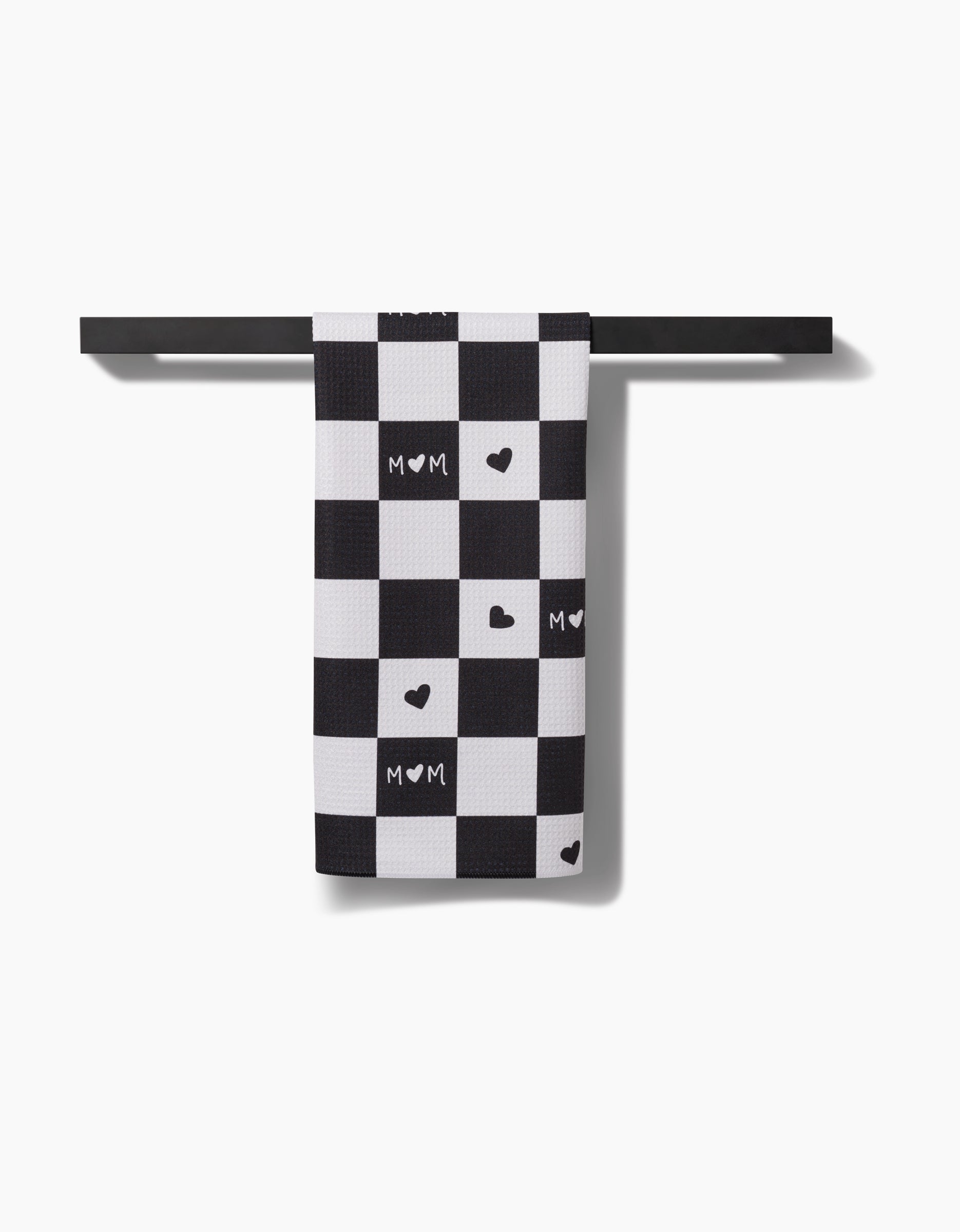 The GEOMETRY Loving Tea Towel hangs on a horizontal bar, showcasing a black and white checkered pattern with heart shapes and MOM on some squares—crafted from durable, stylish Geoweave fabric.