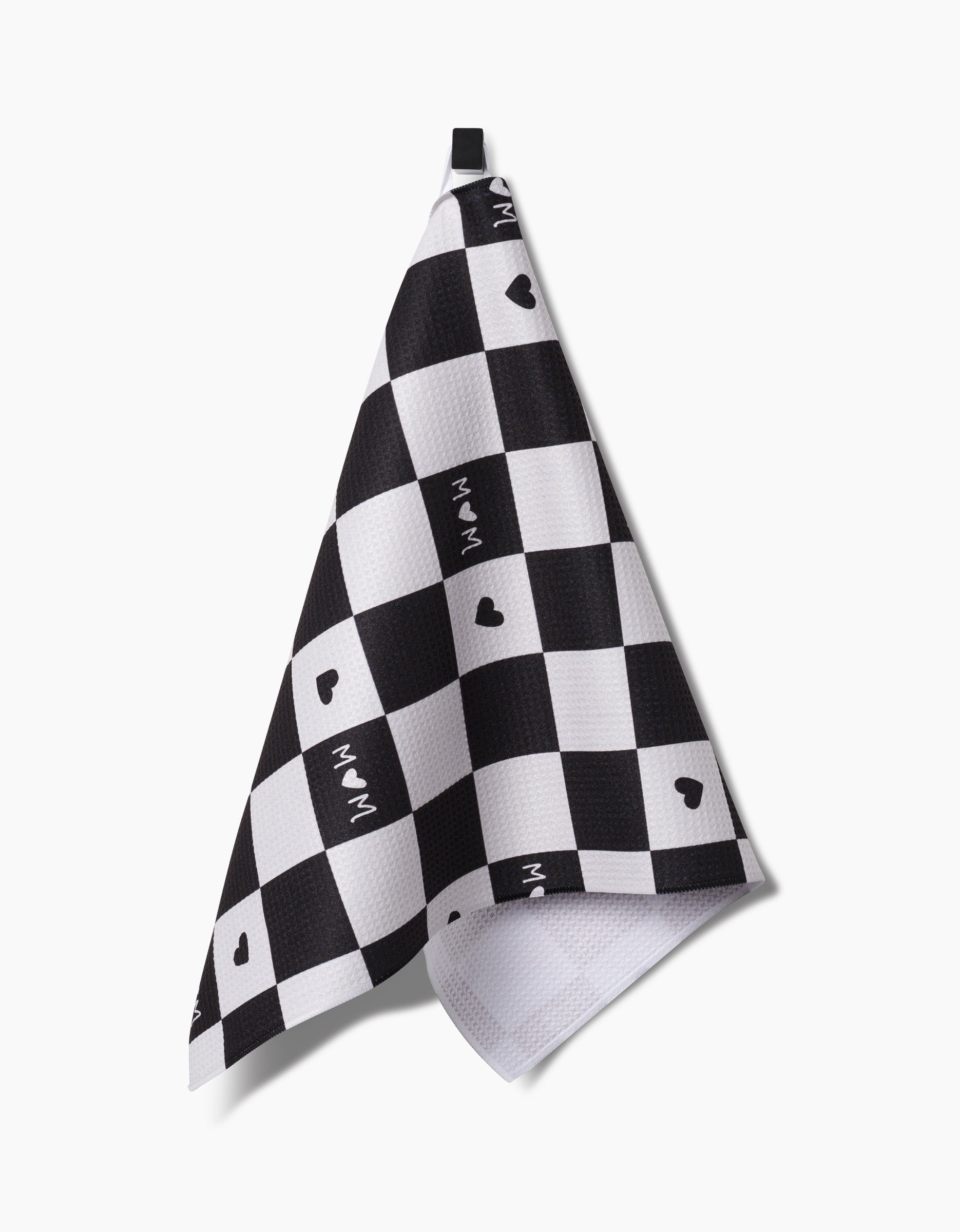 A black and white checkered GEOMETRY Loving Tea Towel hangs on a hook, displaying small black hearts and WAM on some white squares, crafted from durable Geoweave fabric.