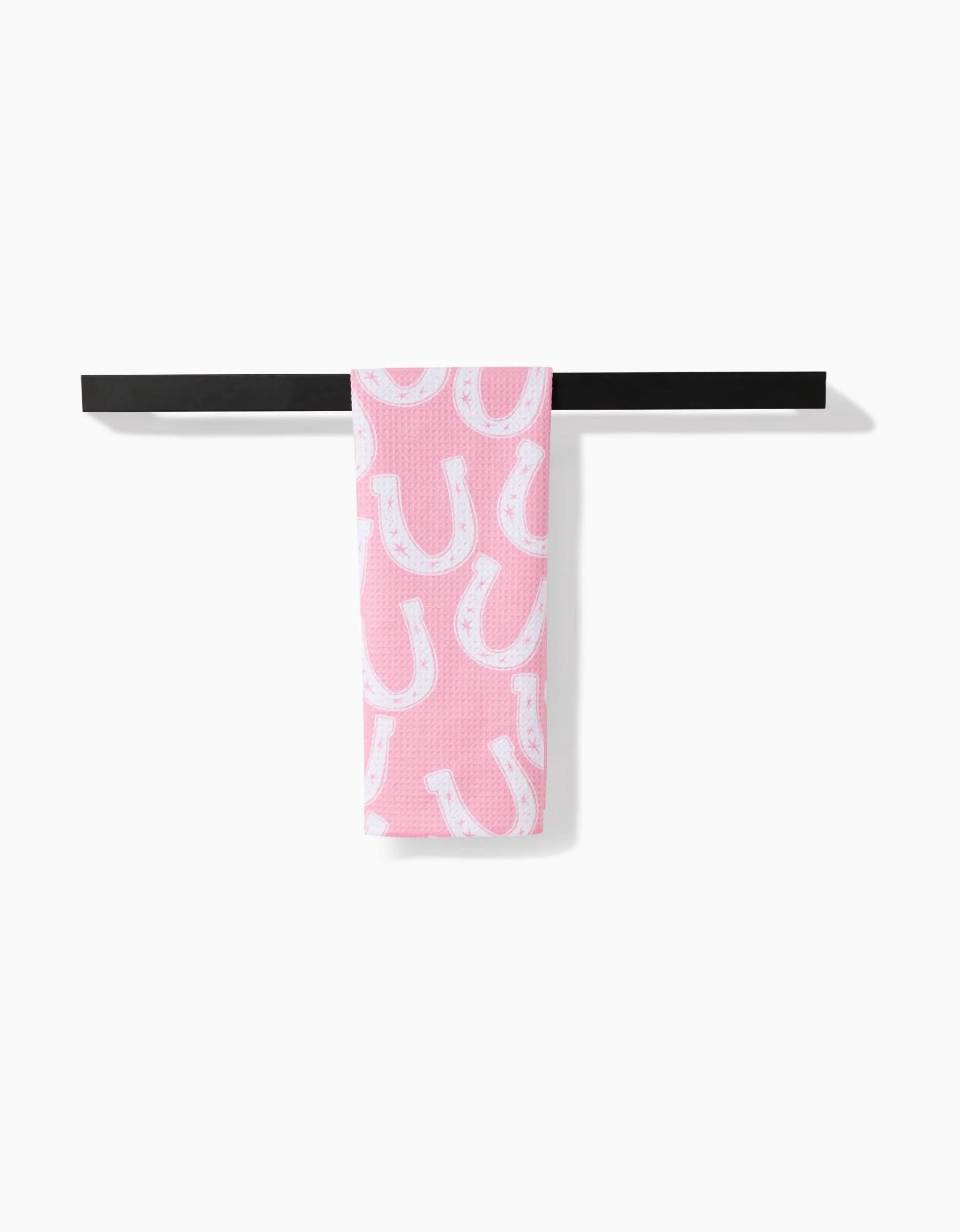The Aubrey Fairchild Lucky Horseshoe tea towel, featuring a white horseshoe pattern on pink Geoweave fabric, is folded over a horizontal black bar against a white background.